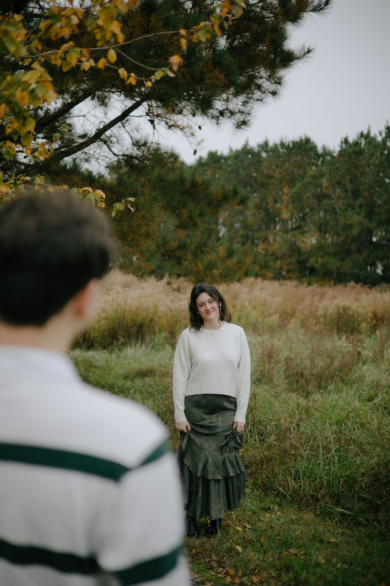 A romantic autumn engagement photo session in a field with golden leaves and evergreen trees in the background.