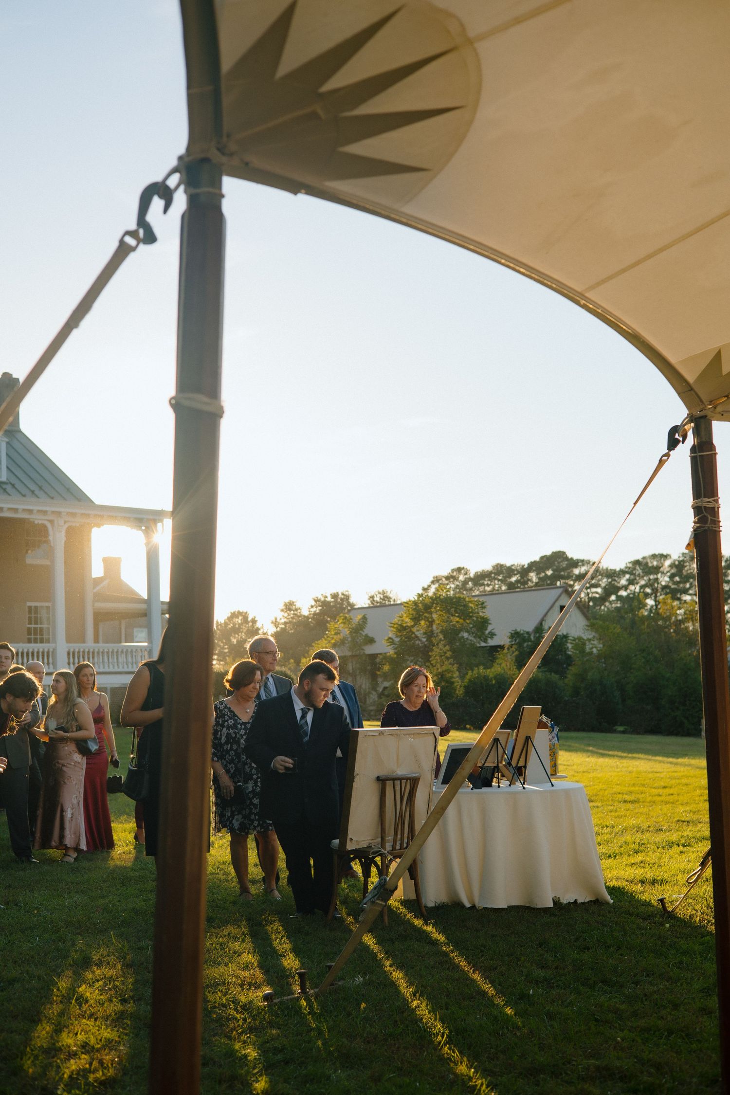 Wedding guests gather under an outdoor tent canopy at sunset with tables and golden light streaming across the lawn.