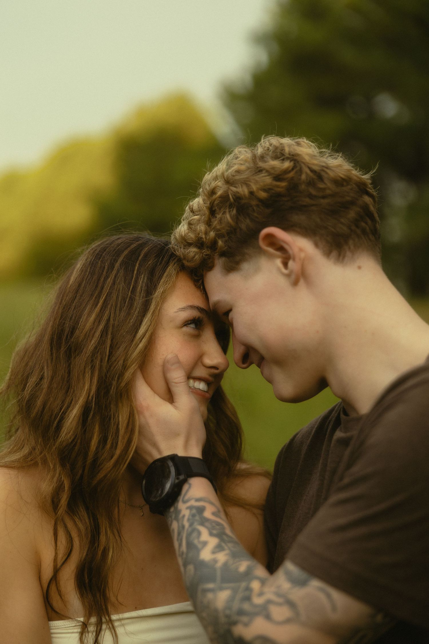 A young couple shares a romantic forehead-to-forehead moment outdoors, with the man's tattooed hand gently cupping her face.