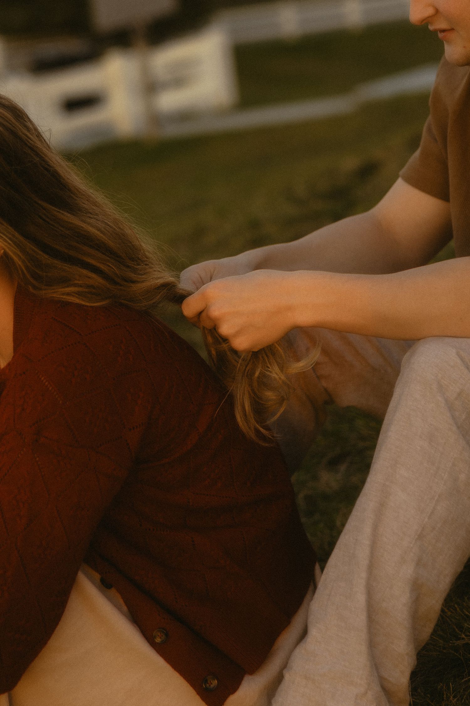 Person braiding another person's long hair outdoors on grass during golden hour sunset light.