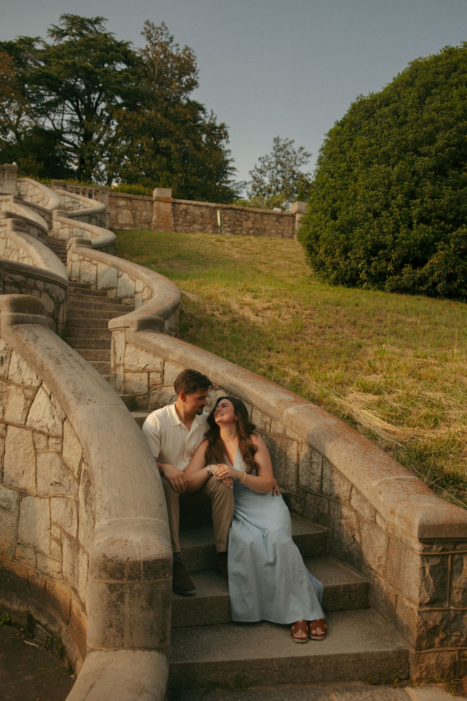 Couple sitting on stone steps in a park, woman in blue dress gazing up at man in warm golden light.