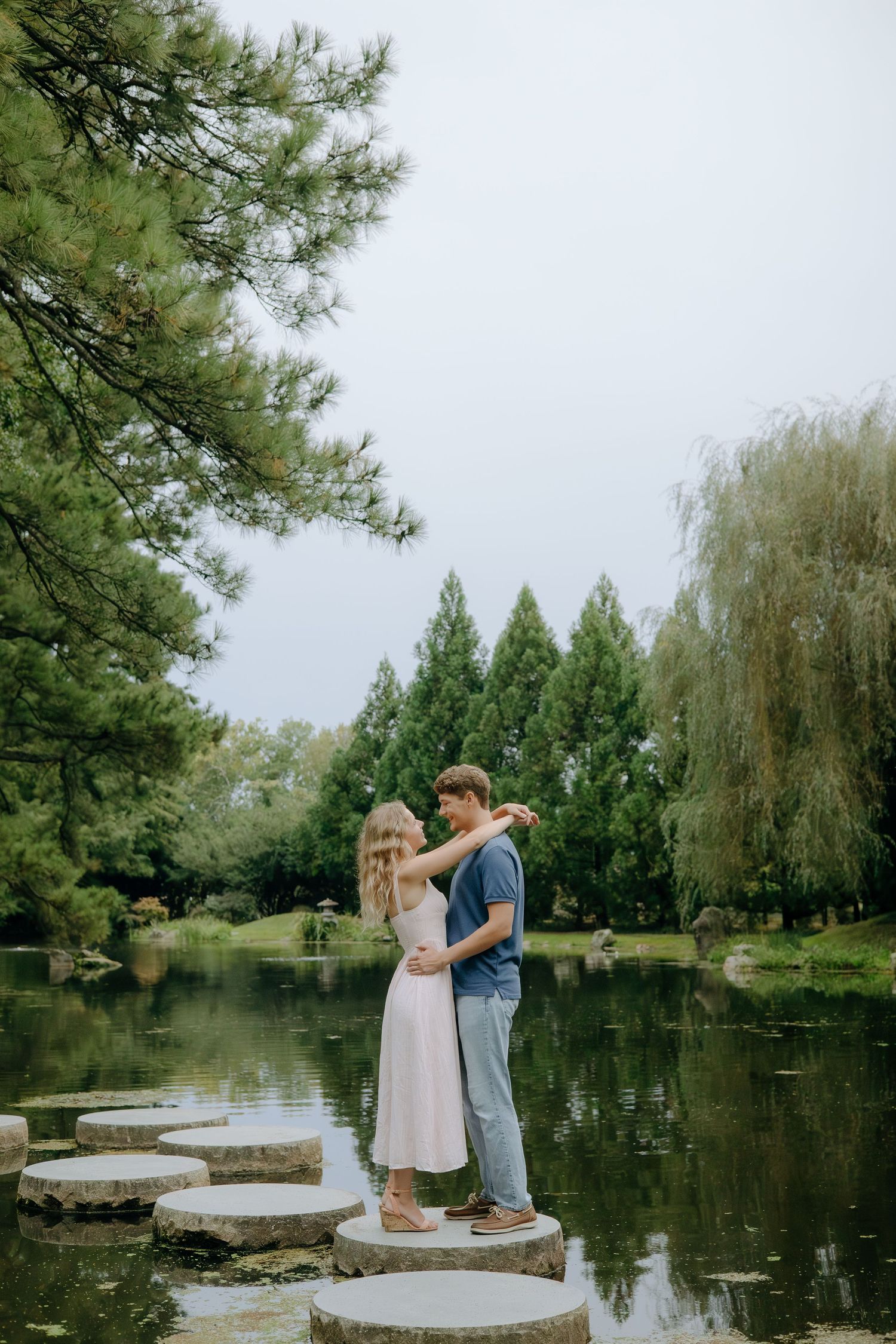 Couple standing on stepping stones over a tranquil pond surrounded by lush green trees in a serene garden setting.