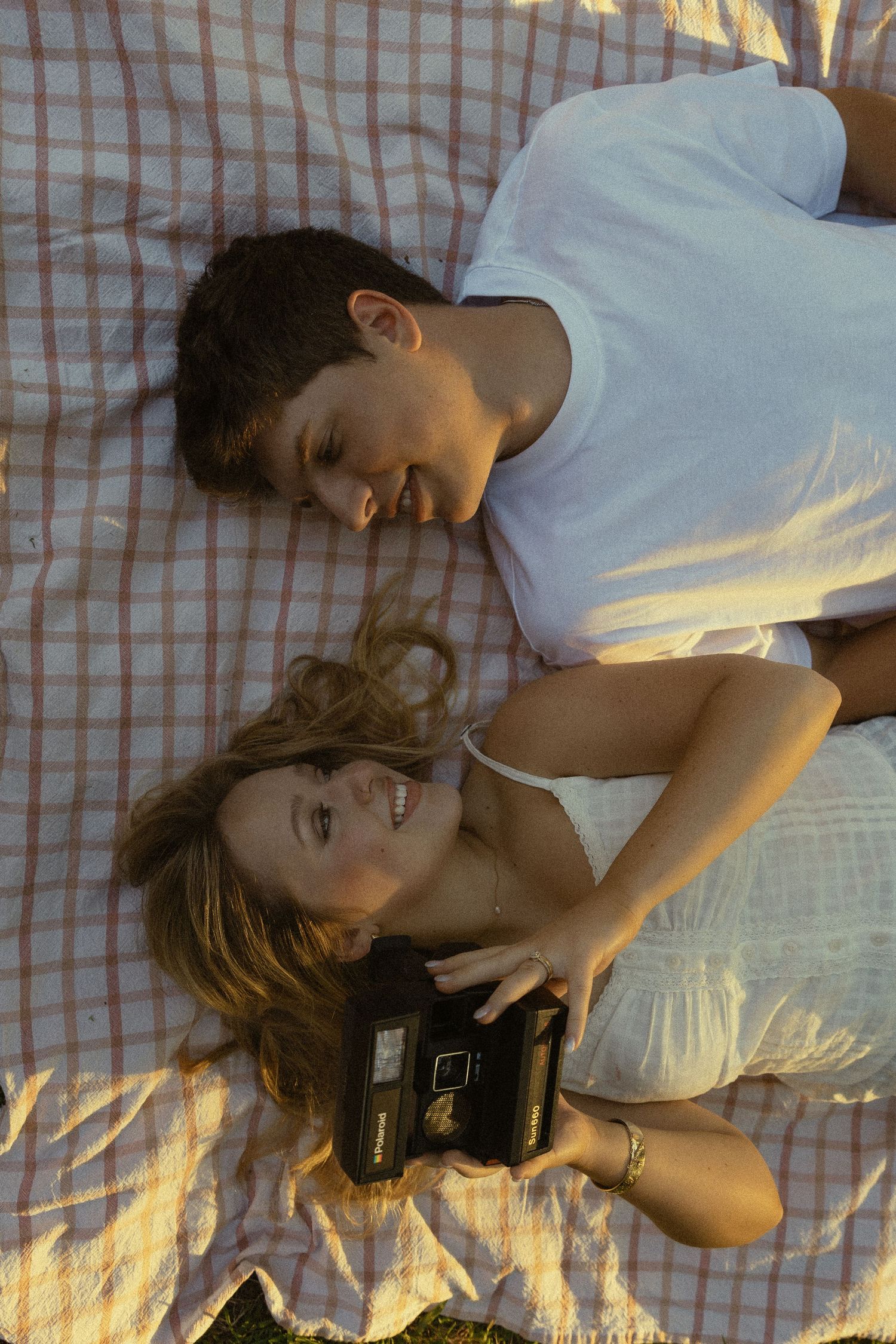Couple lying on a picnic blanket outdoors, smiling at each other while holding a vintage camera.