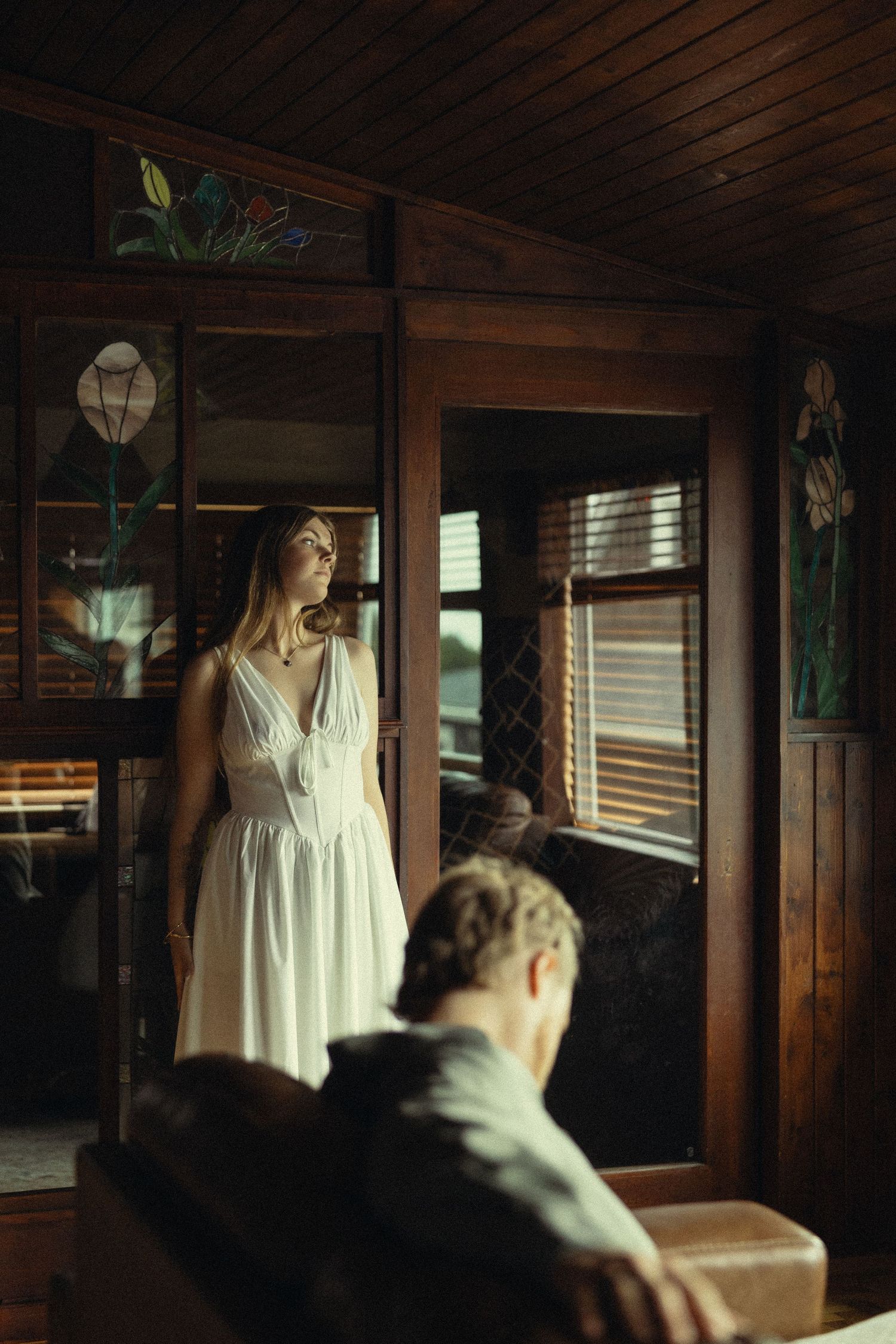 A woman in a white dress stands in a rustic wood-paneled room with stained glass windows while a man sits in the foreground.