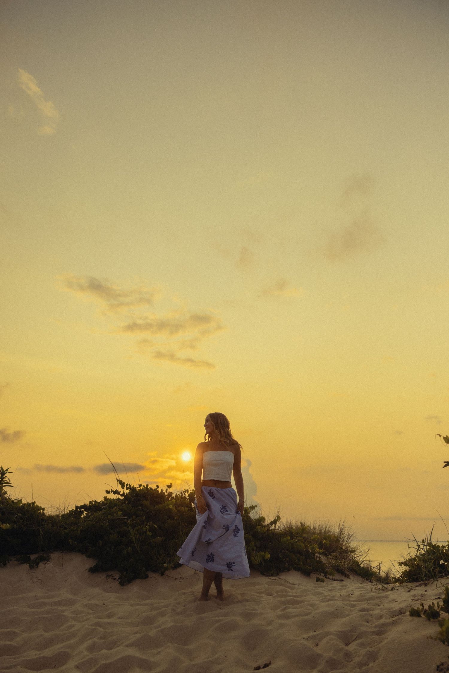 Woman in white crop top and floral skirt standing on sandy dune watching golden sunset over ocean.