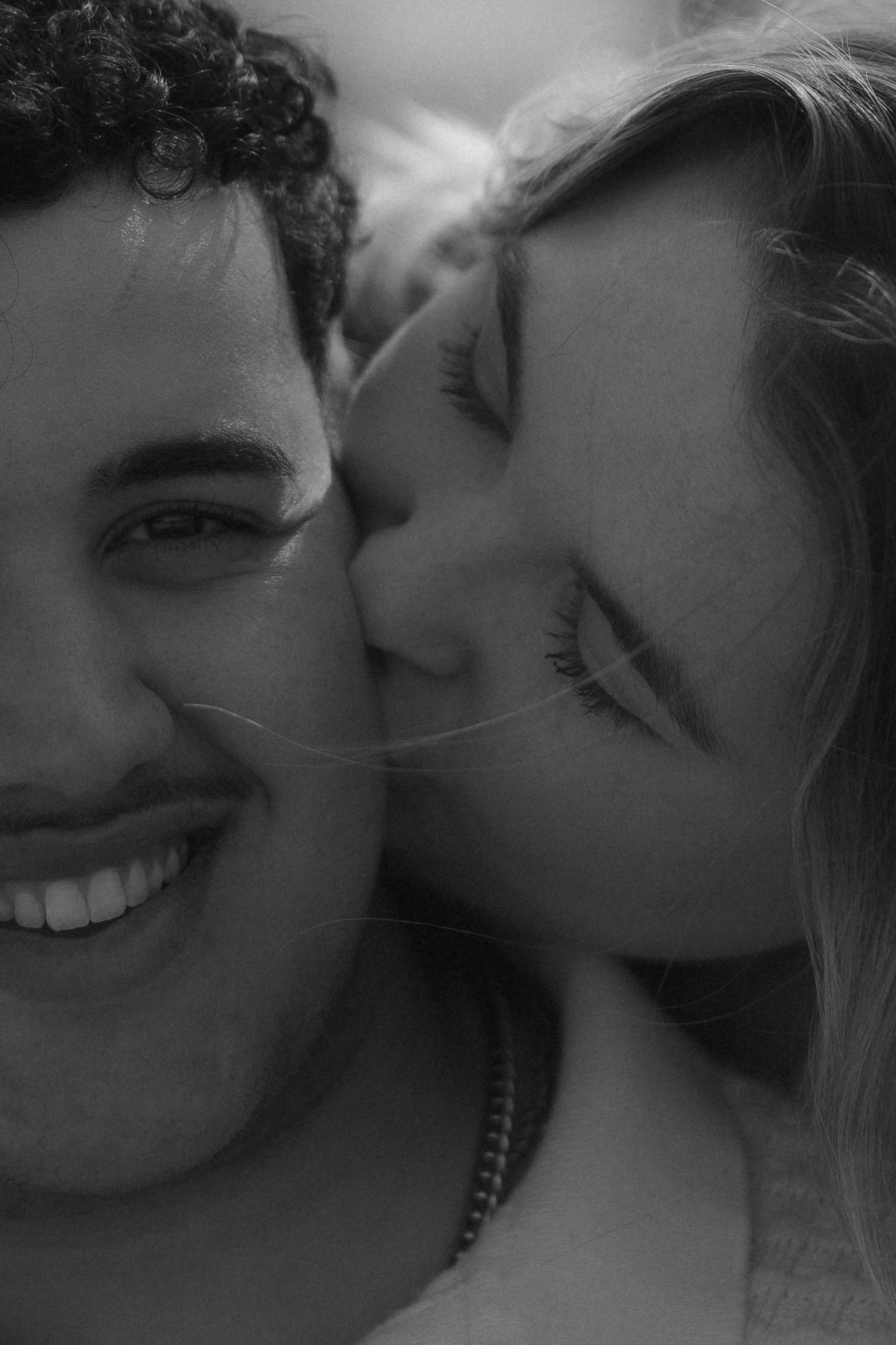 Black and white close-up of a smiling man being kissed on the cheek by a blonde woman.
