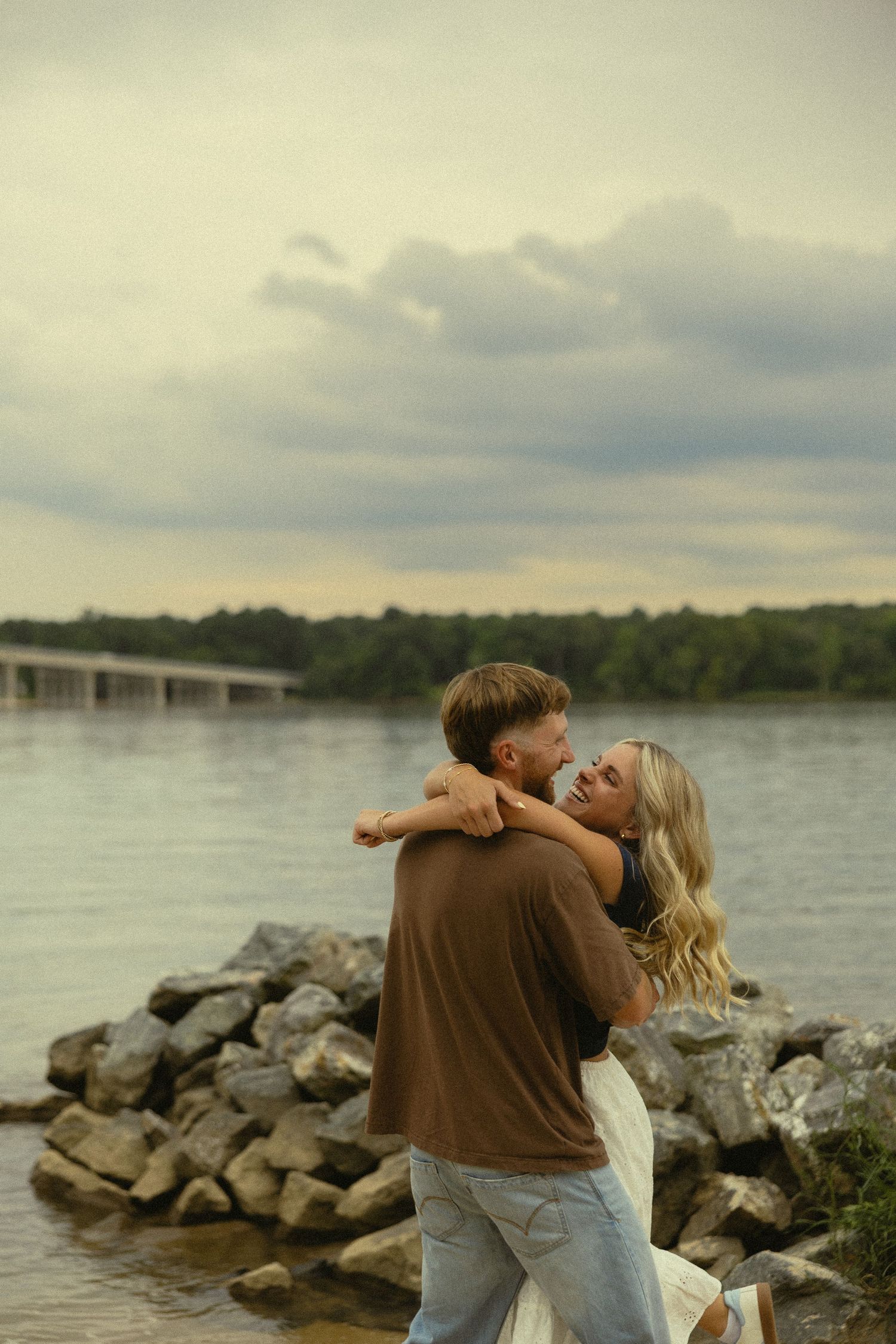 Couple embracing romantically by a rocky riverbank at dusk, with a bridge and lush trees in the background.