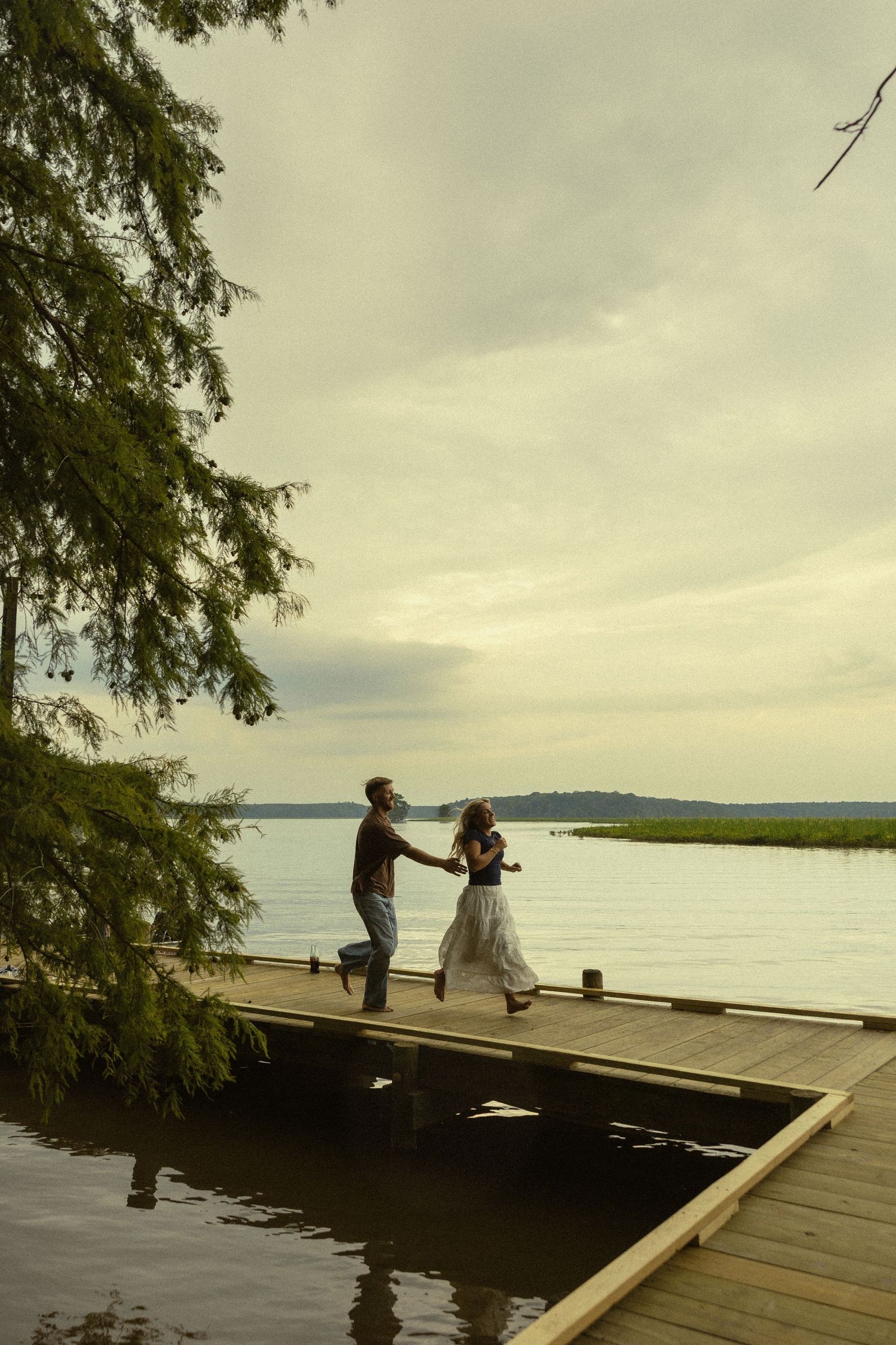 Couple holding hands and dancing on a wooden dock by a serene lake at dusk, surrounded by lush green trees.