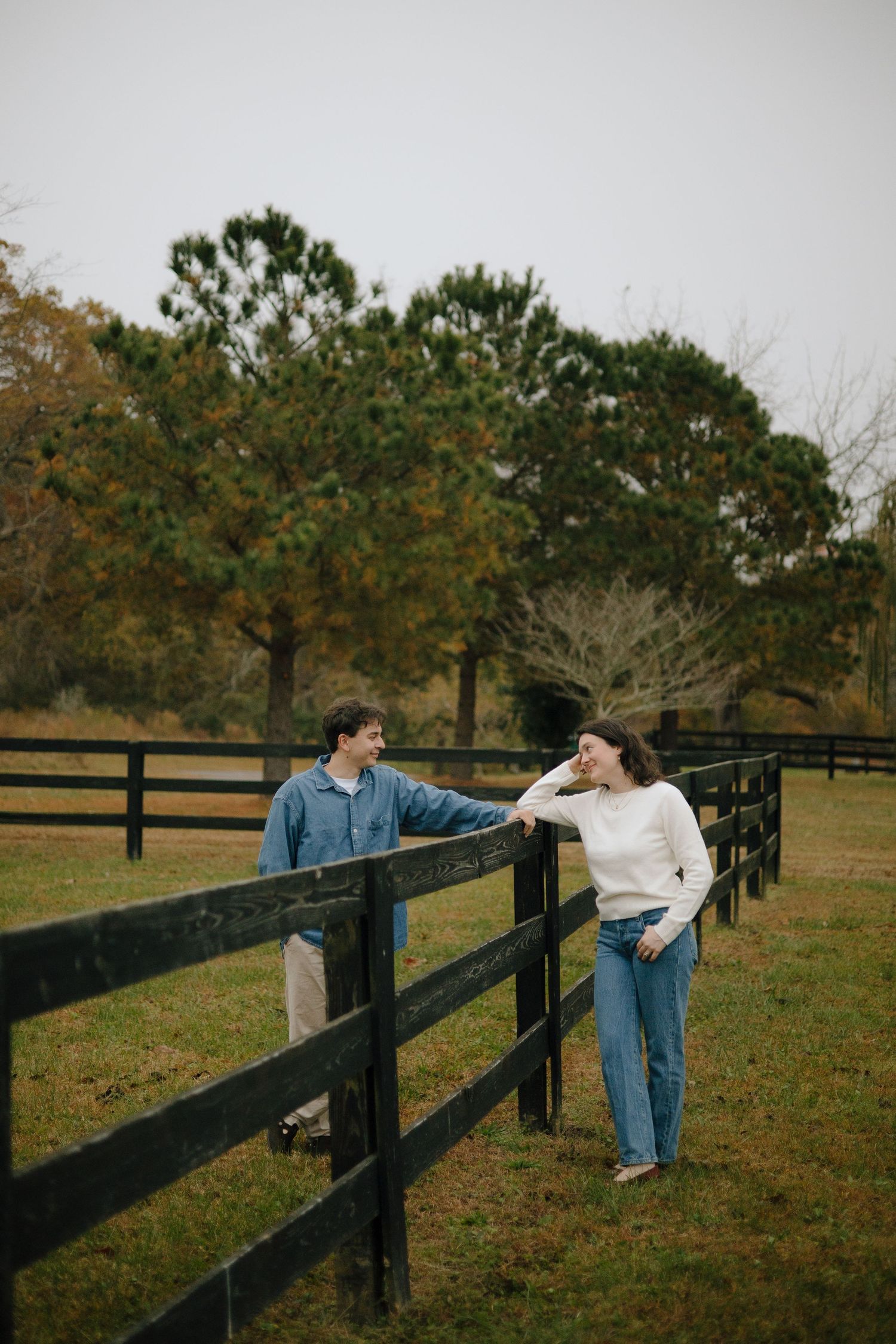 Couple in casual attire posing along a black fence in an autumn setting.