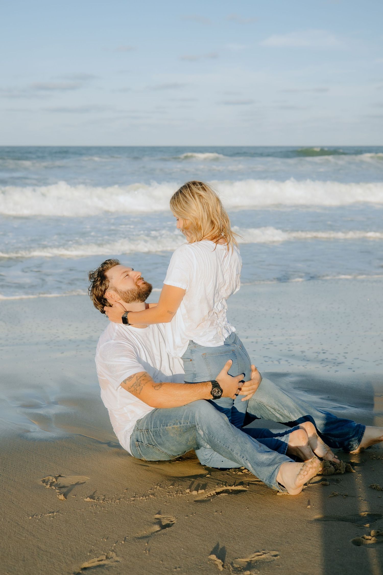 A couple plays and laughs while sitting on the sandy beach near the ocean.