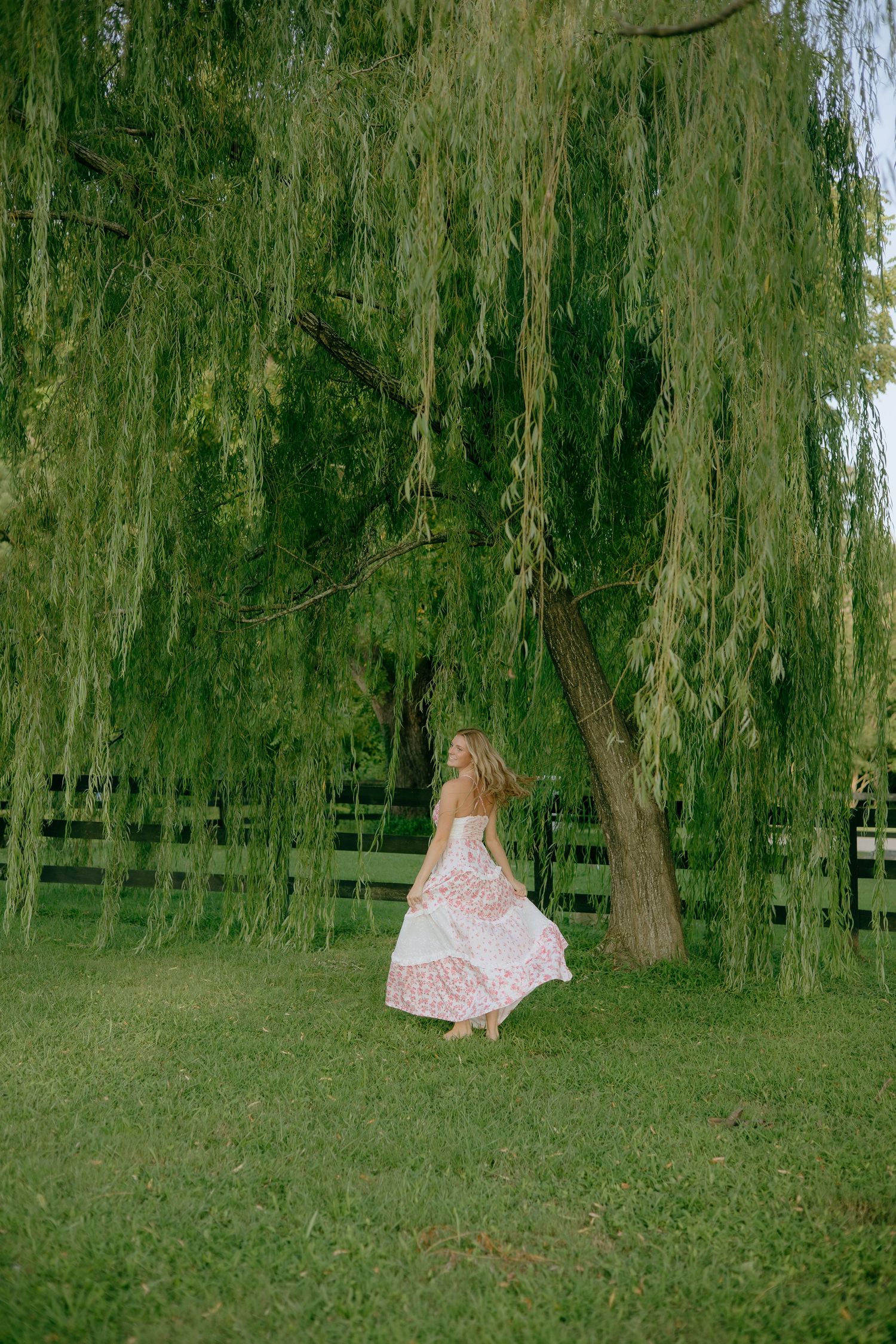 A figure in a flowy pink dress twirls gracefully beneath drooping willow trees in a serene garden setting.