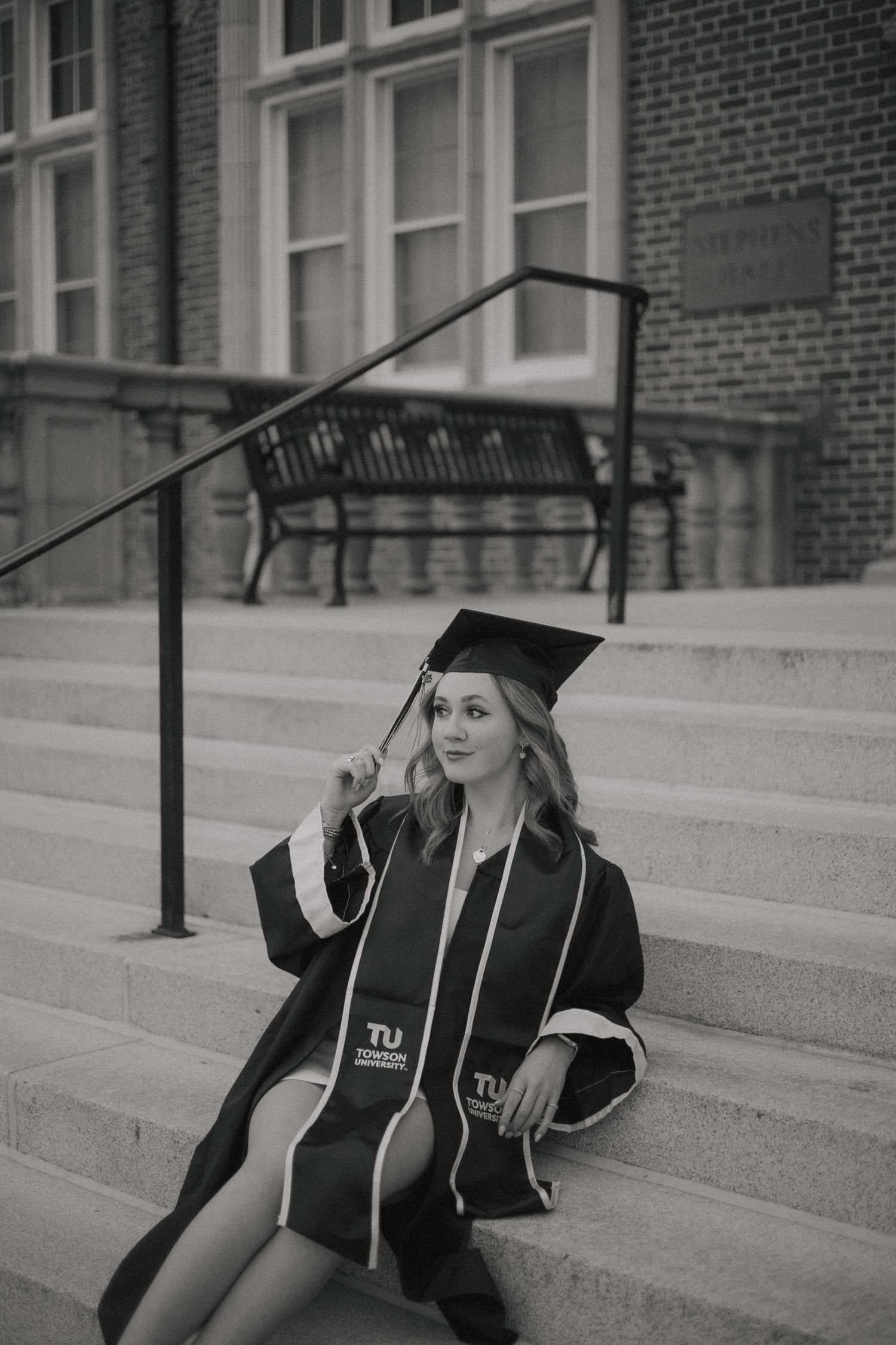 Graduate in cap and gown sitting on stone steps of brick building in black and white photograph.
