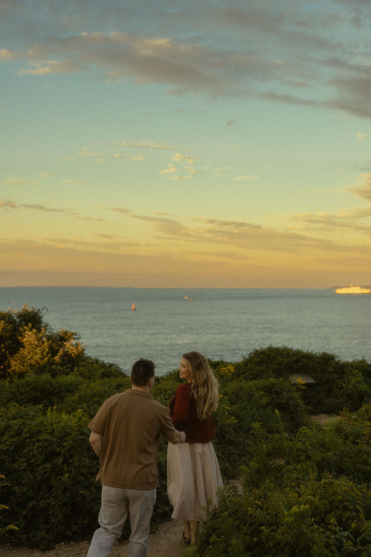 Couple standing together overlooking a scenic ocean sunset surrounded by lush green bushes at golden hour.