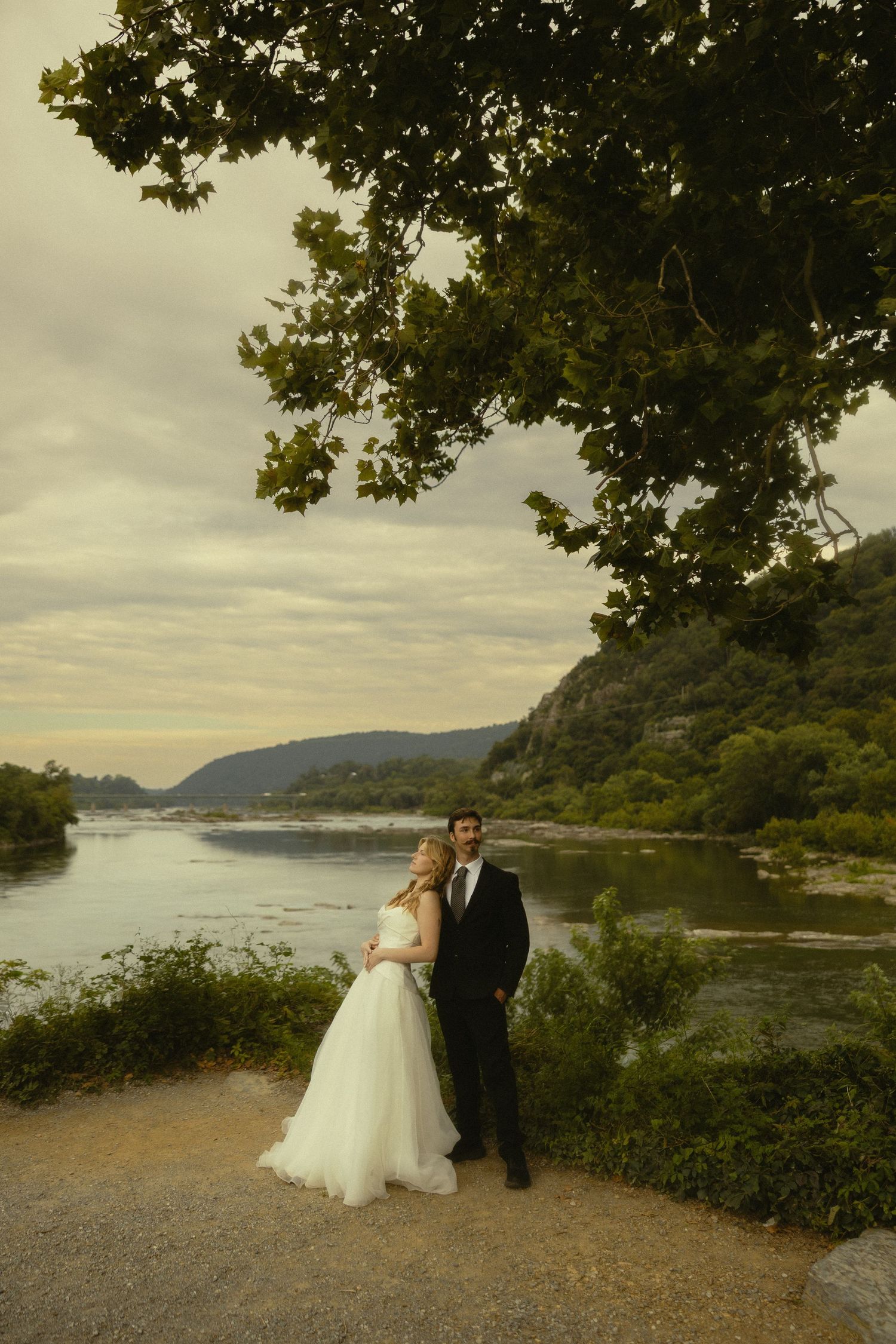 Bride and groom posing by a scenic river with lush green hills and a large tree at dusk.