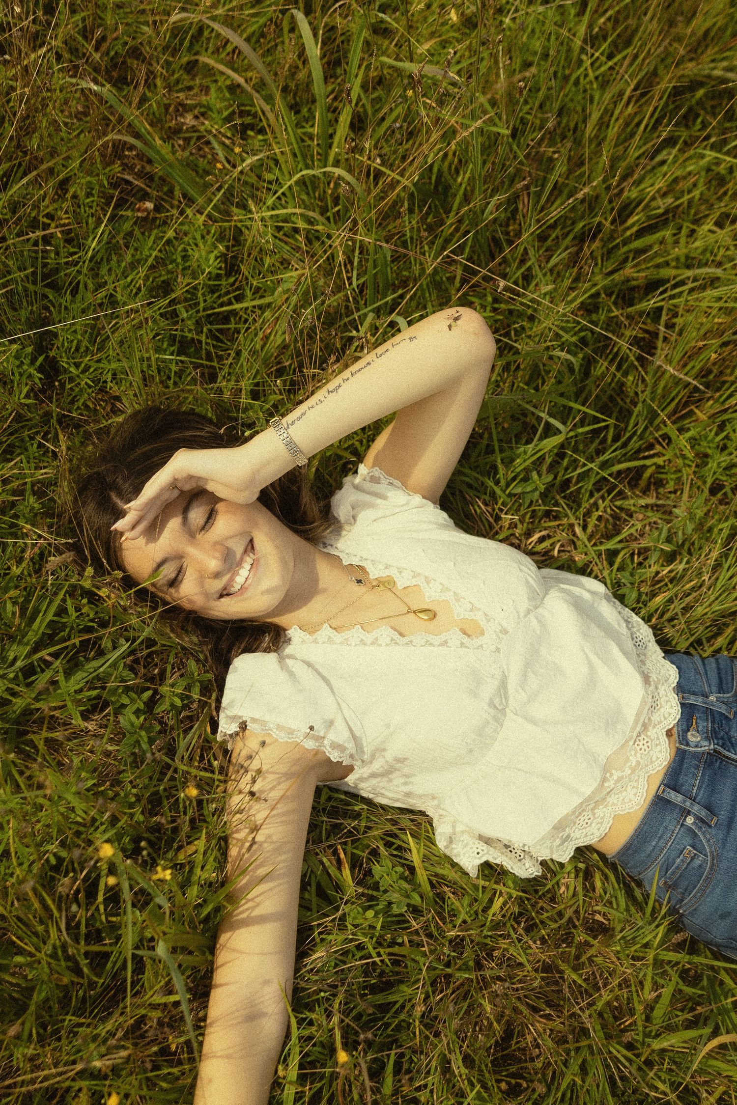 Young woman lying in green grass, smiling with eyes closed, wearing white lace top and denim shorts on sunny day.