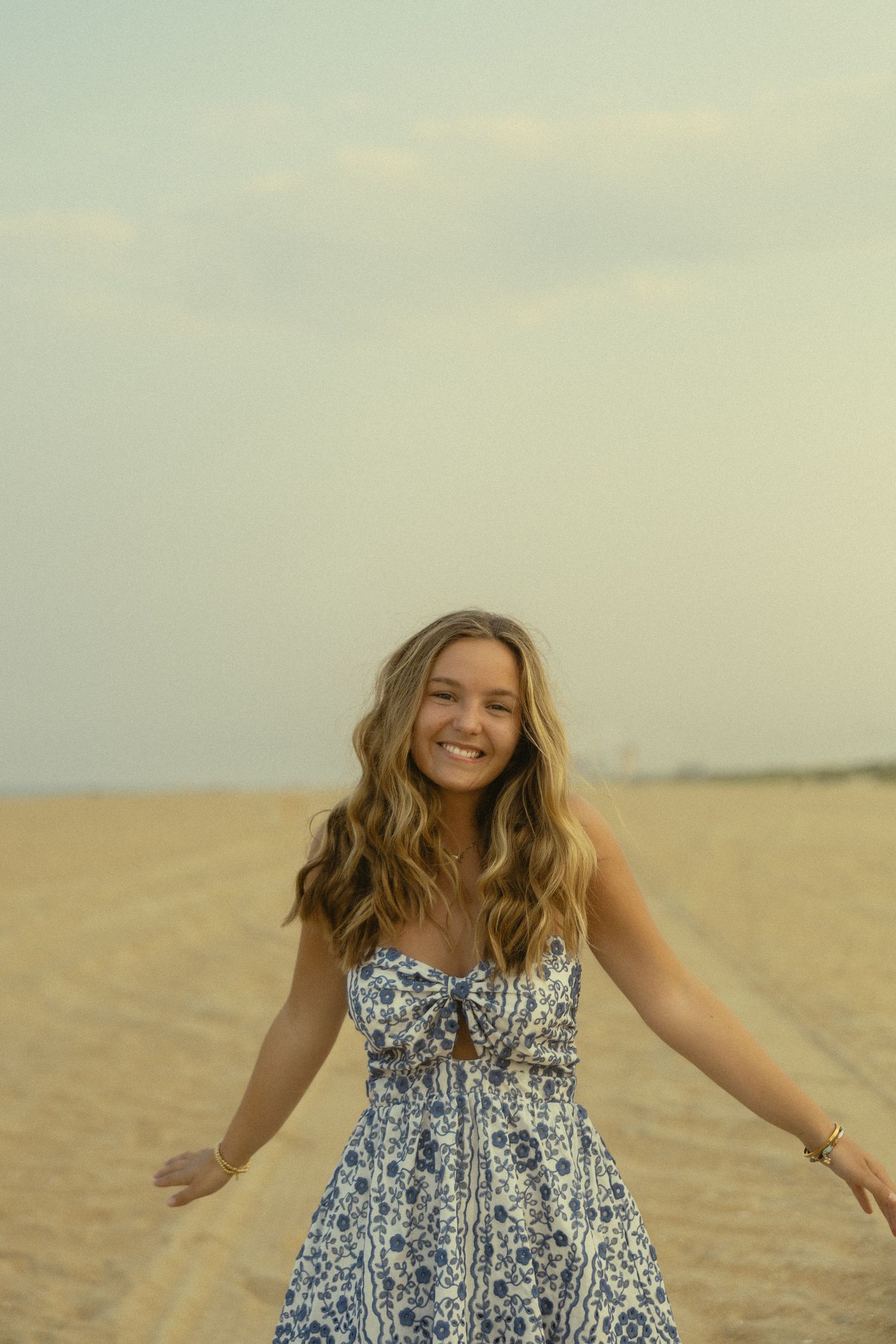Young woman in blue floral sundress smiling on sandy beach with overcast sky.