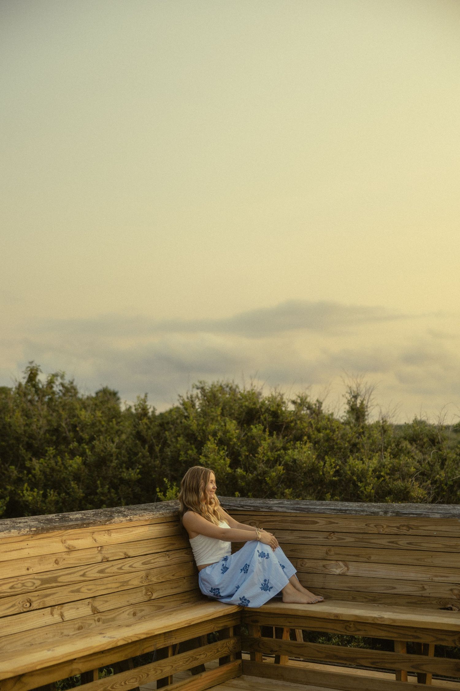 A young woman in a blue floral skirt sits on a wooden deck bench at sunset, gazing at lush greenery.