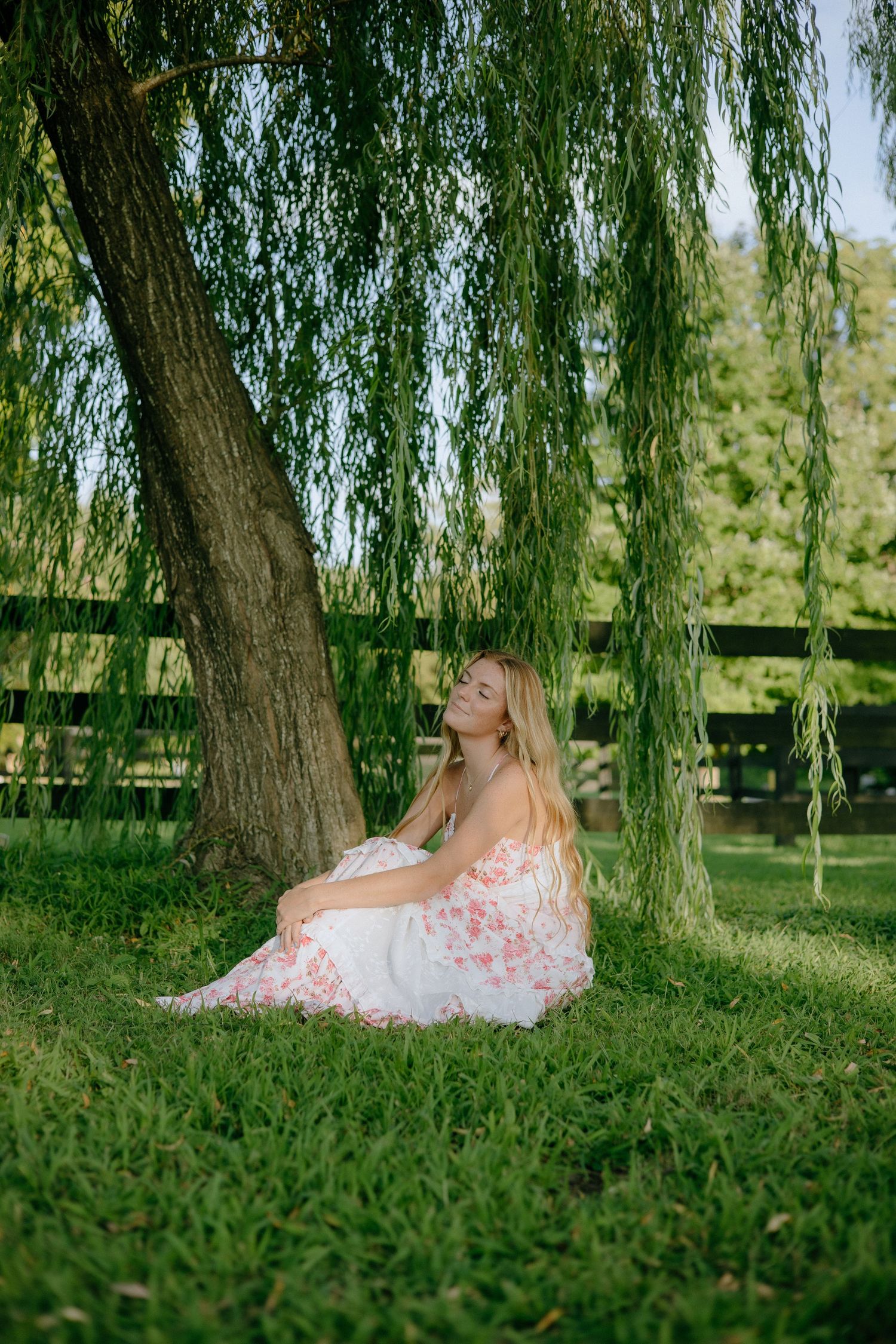 A person in a white lace dress lies serenely under a weeping willow tree in a lush green garden setting.