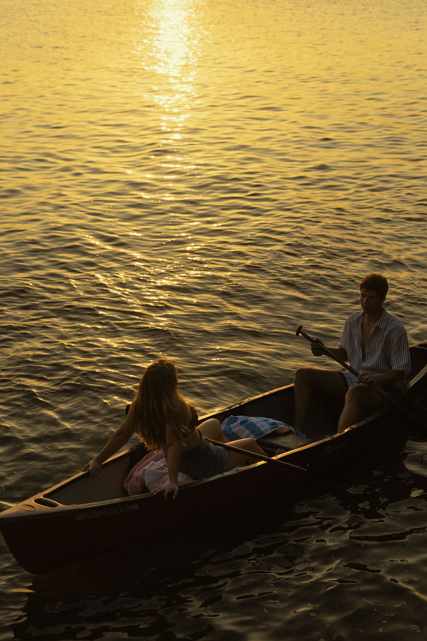 Couple enjoying a romantic sunset canoe ride on golden reflective water, man holding a bottle.