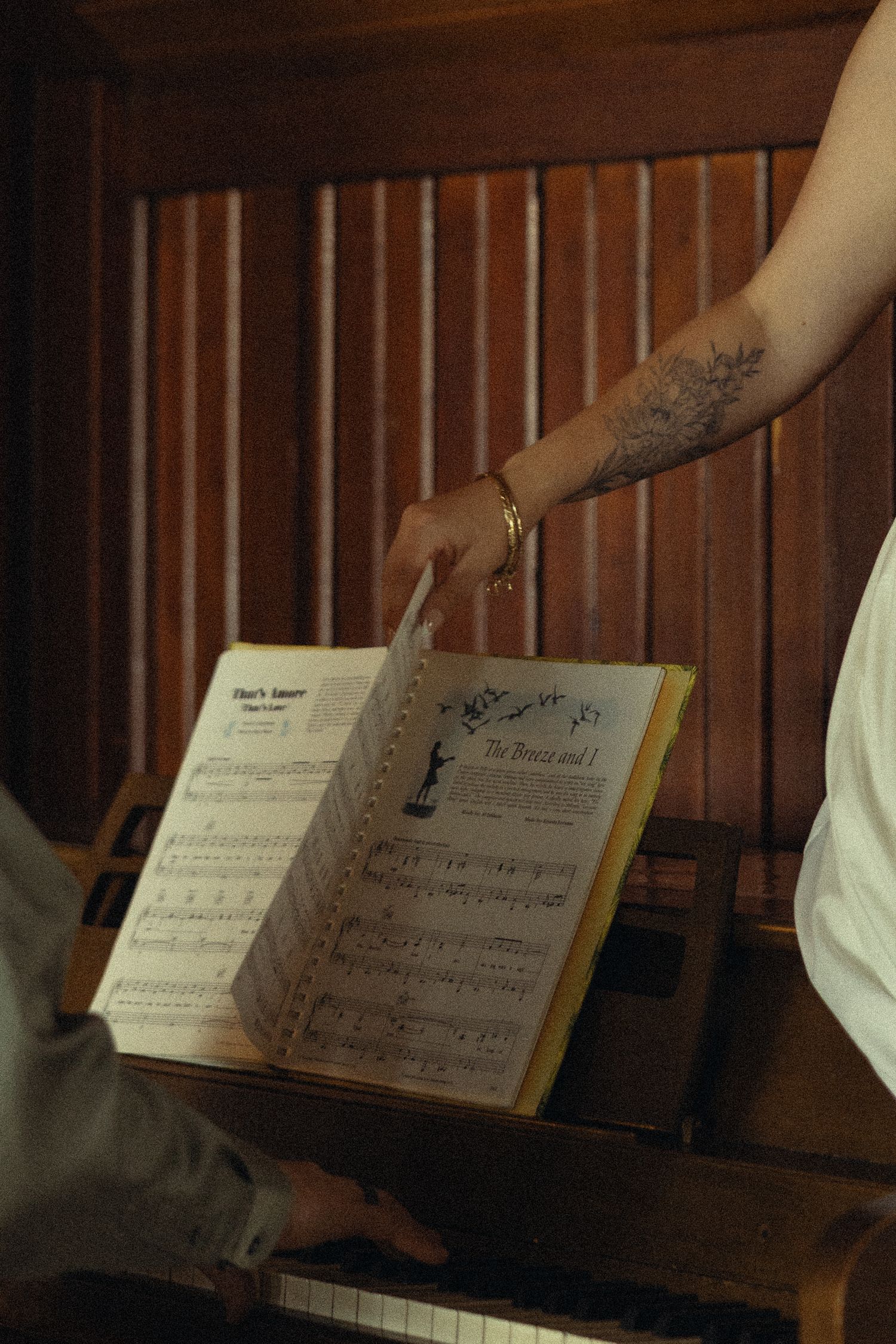 Tattooed hand turning sheet music pages on a vintage piano, with a pianist playing in the background.