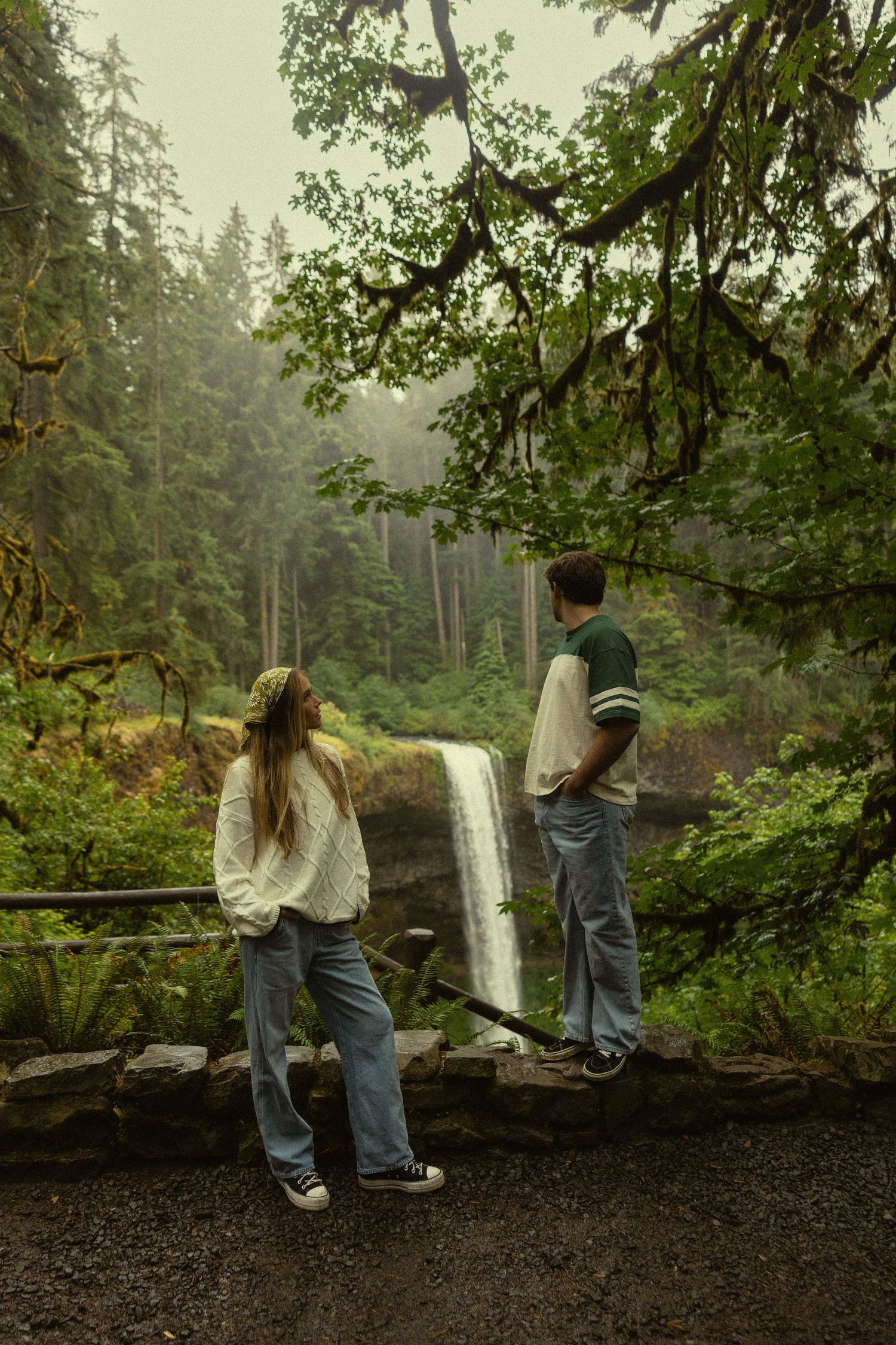 Two people admire a misty waterfall surrounded by lush green forest trees on a foggy day.