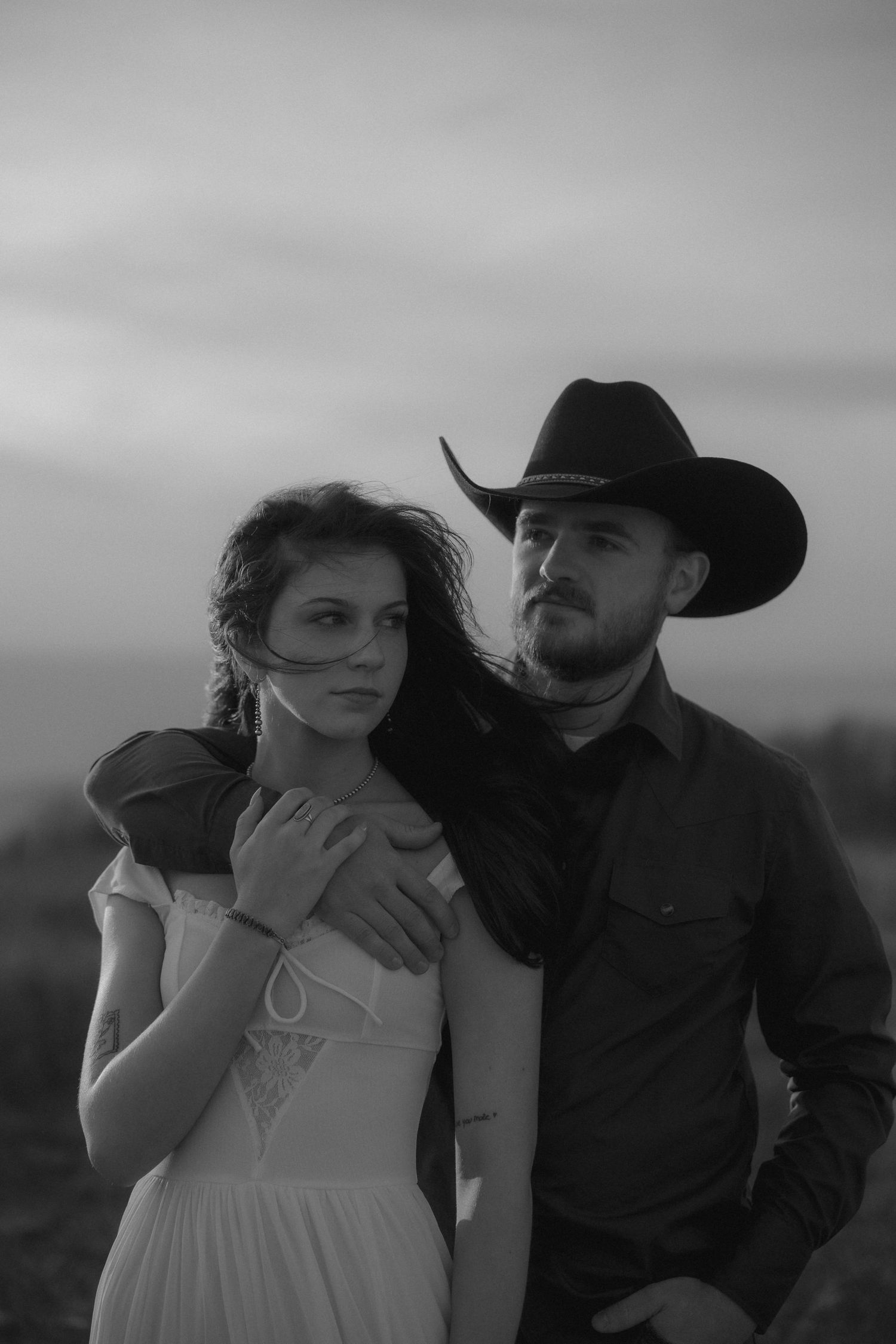 Black and white photo of a couple, man in cowboy hat embracing woman in white dress outdoors at dusk.