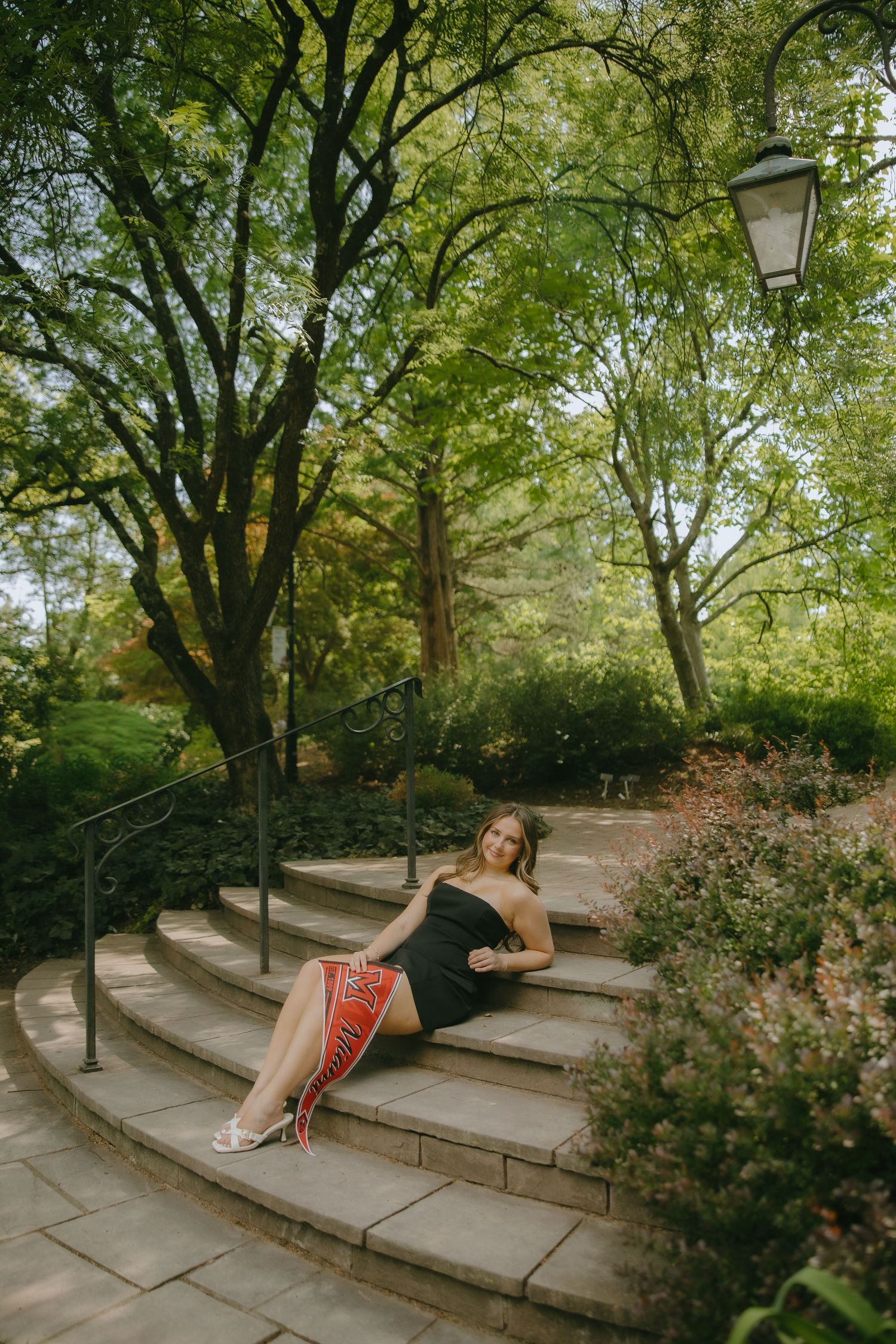 Woman in black dress with red accents sits on curved stone steps surrounded by lush green trees in a park.