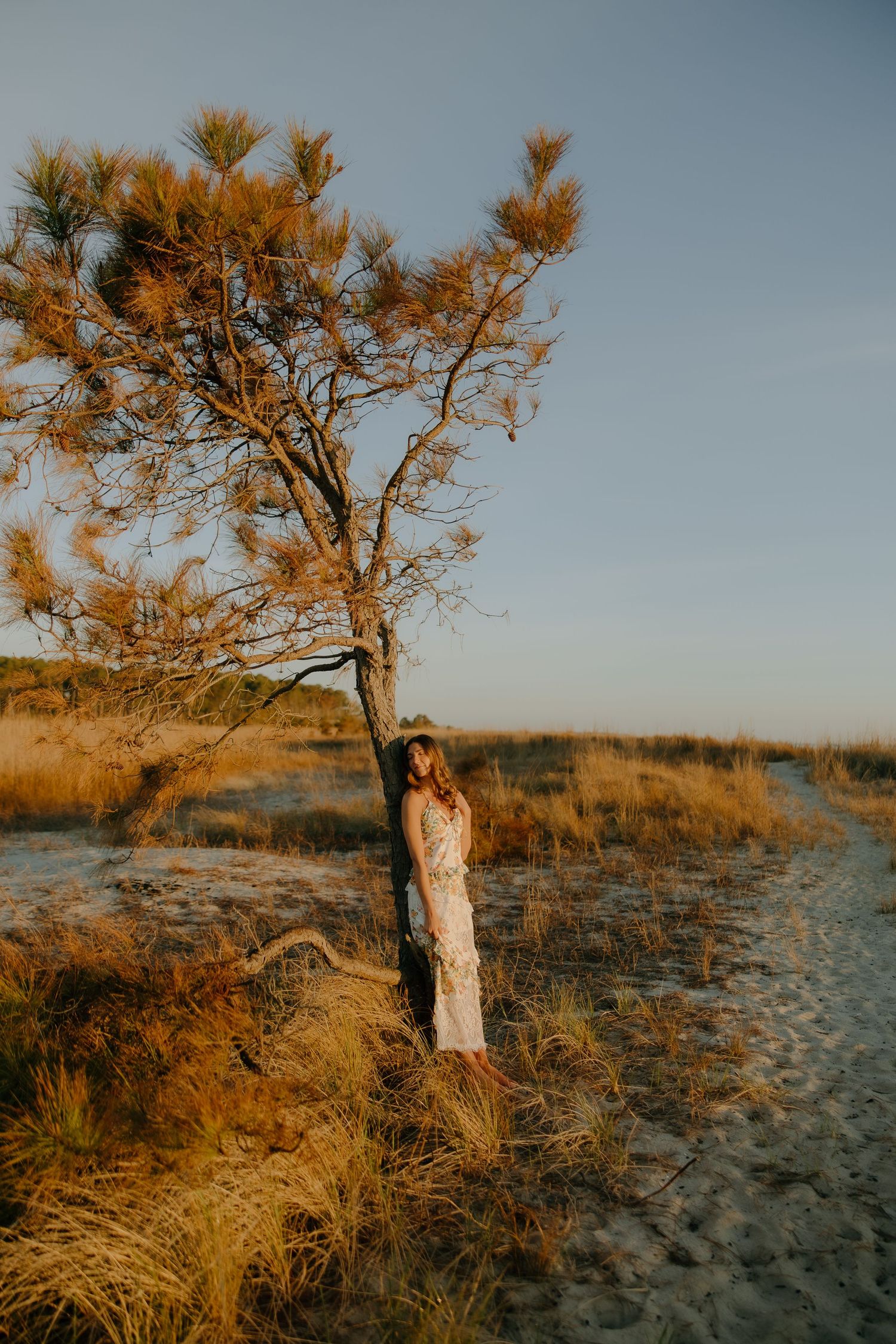 Woman in white dress leaning against a pine tree in golden sandy dunes at sunset.