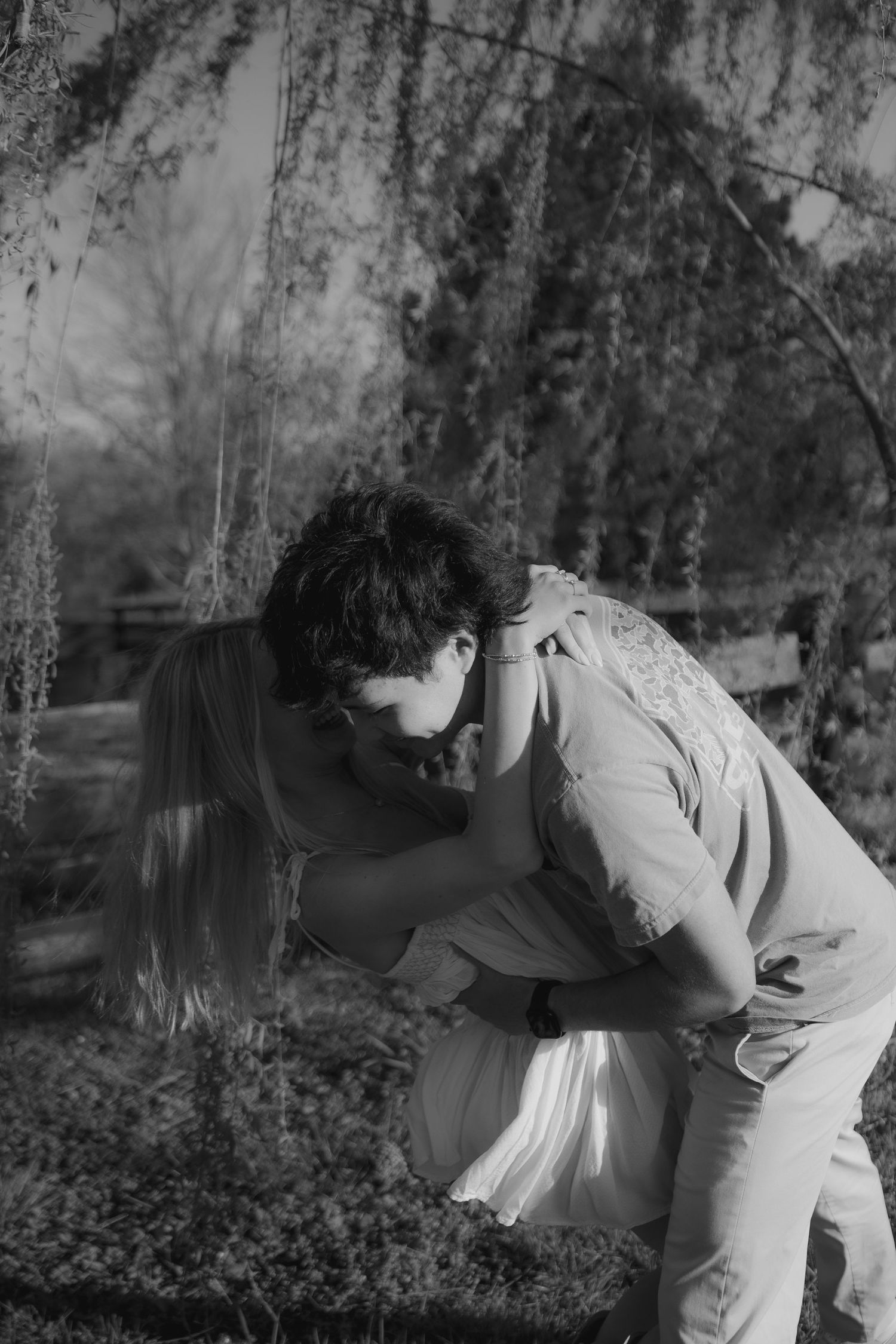 Black and white photo of a couple sharing a romantic embrace under a weeping willow tree outdoors.