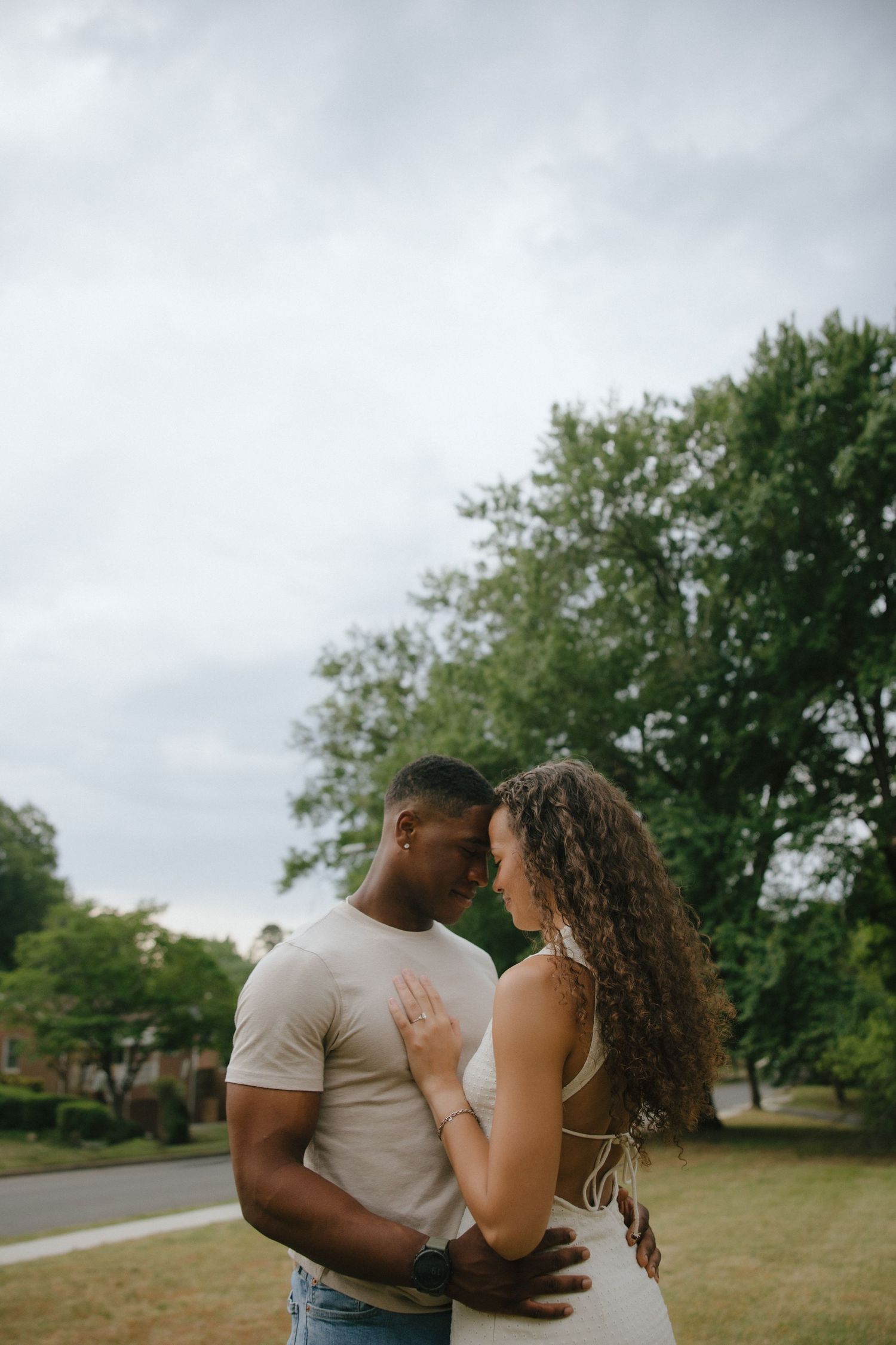 A couple shares an intimate moment outdoors, touching foreheads while embracing in a lush green park under a cloudy sky.