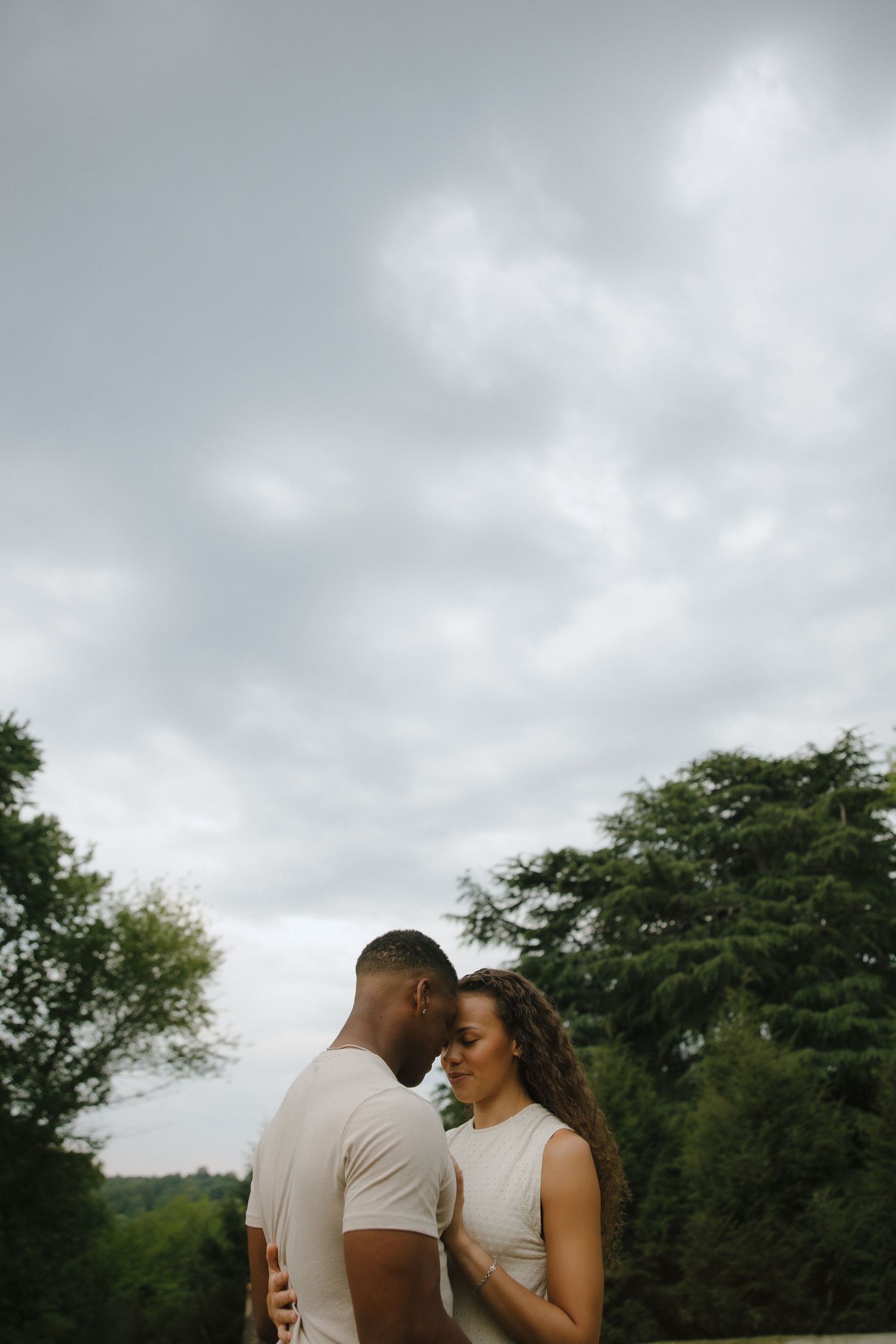 Romantic couple in beige outfits sharing an intimate moment outdoors with lush green trees and cloudy sky backdrop.