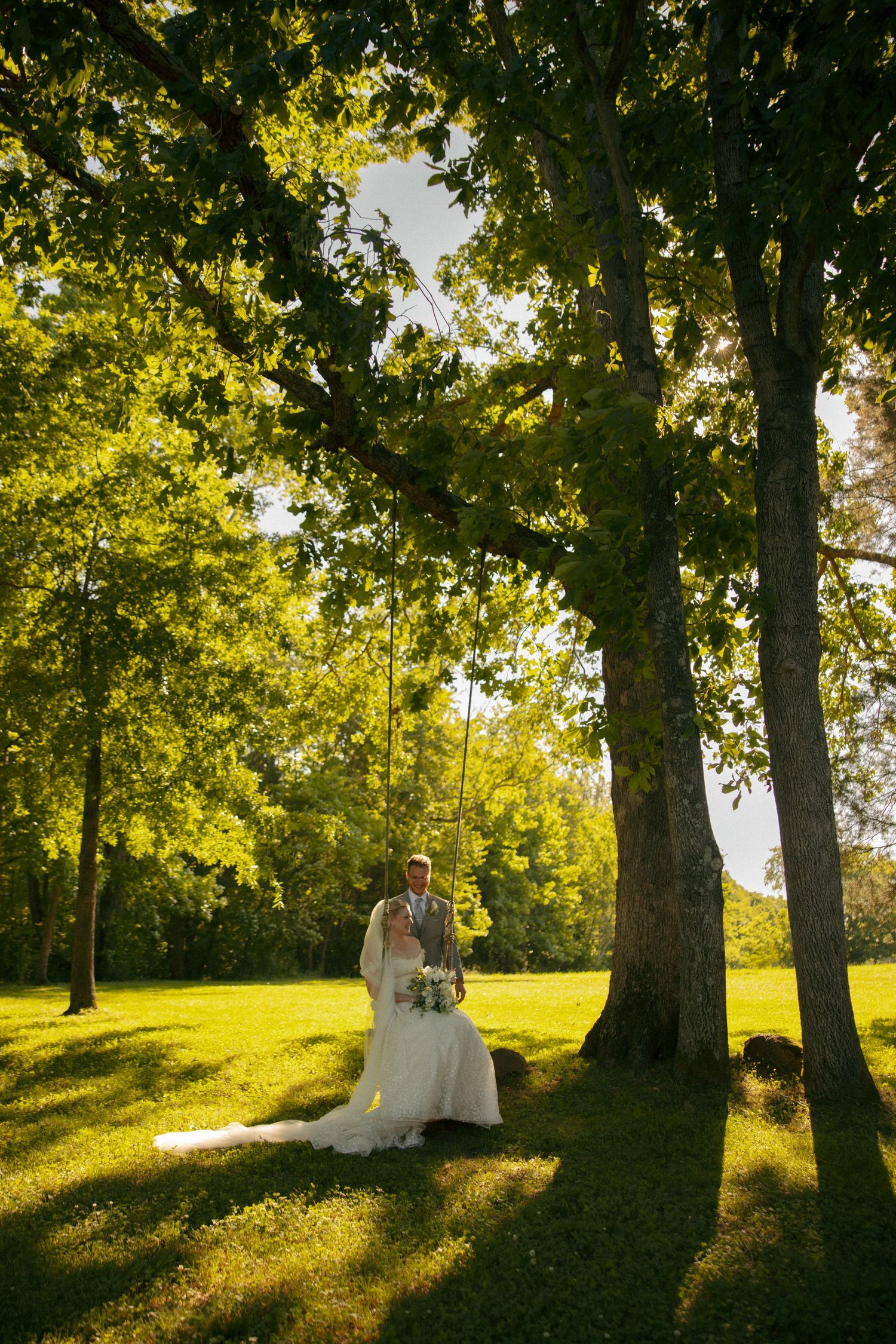 Bride and groom stand beneath sunlit trees on lush green grass, bride holding bouquet in elegant white gown at Avonlea Farms in Mineral, VA