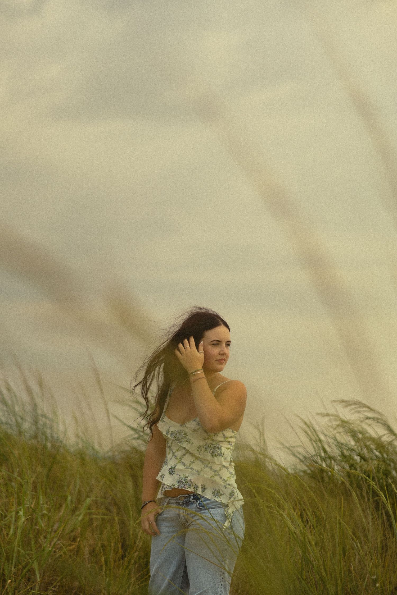 Young woman in floral top and jeans standing in tall grass field with wind blowing her hair on cloudy day.