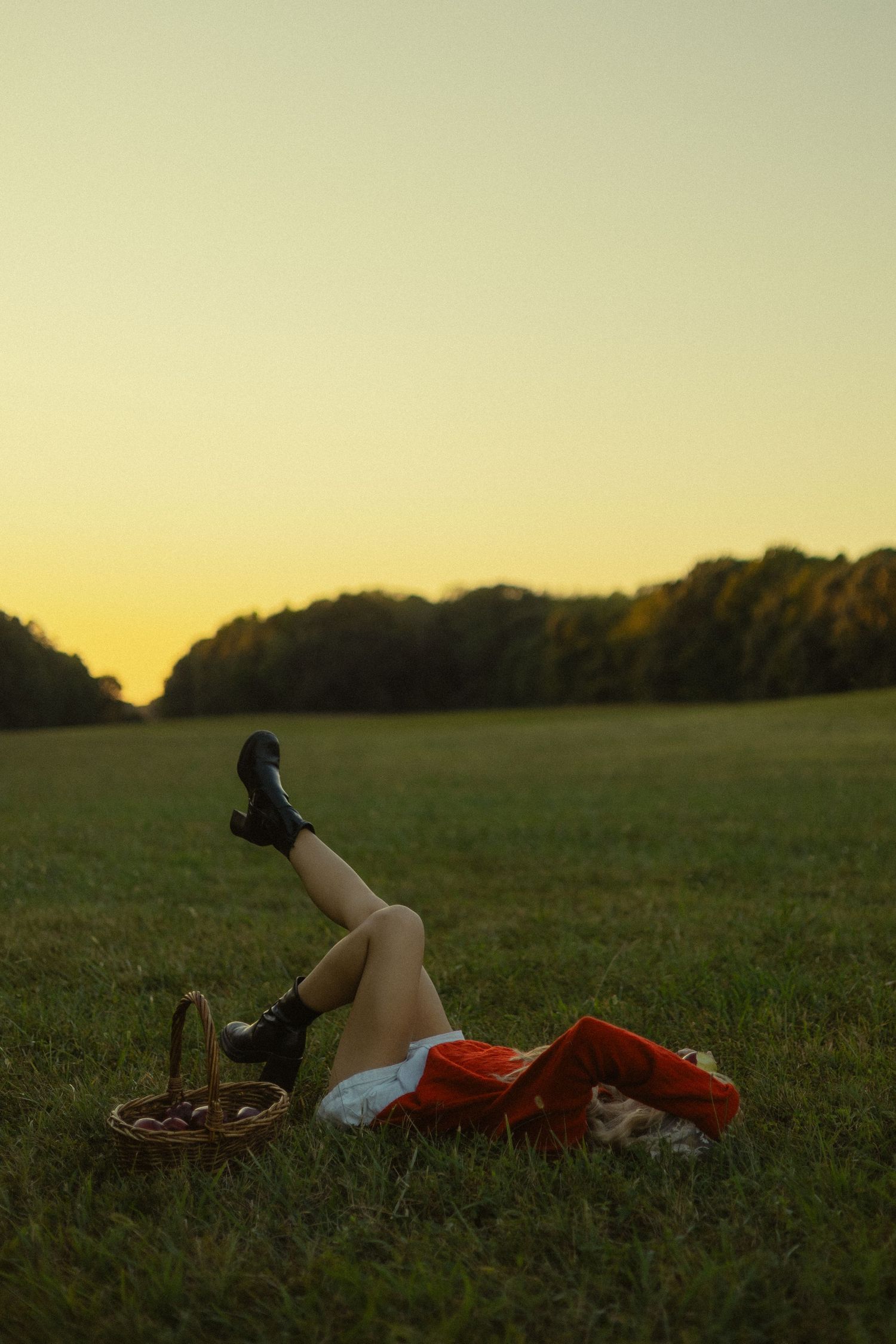 Woman in red skirt and black boots lying on green grass field at sunset with a picnic basket nearby.