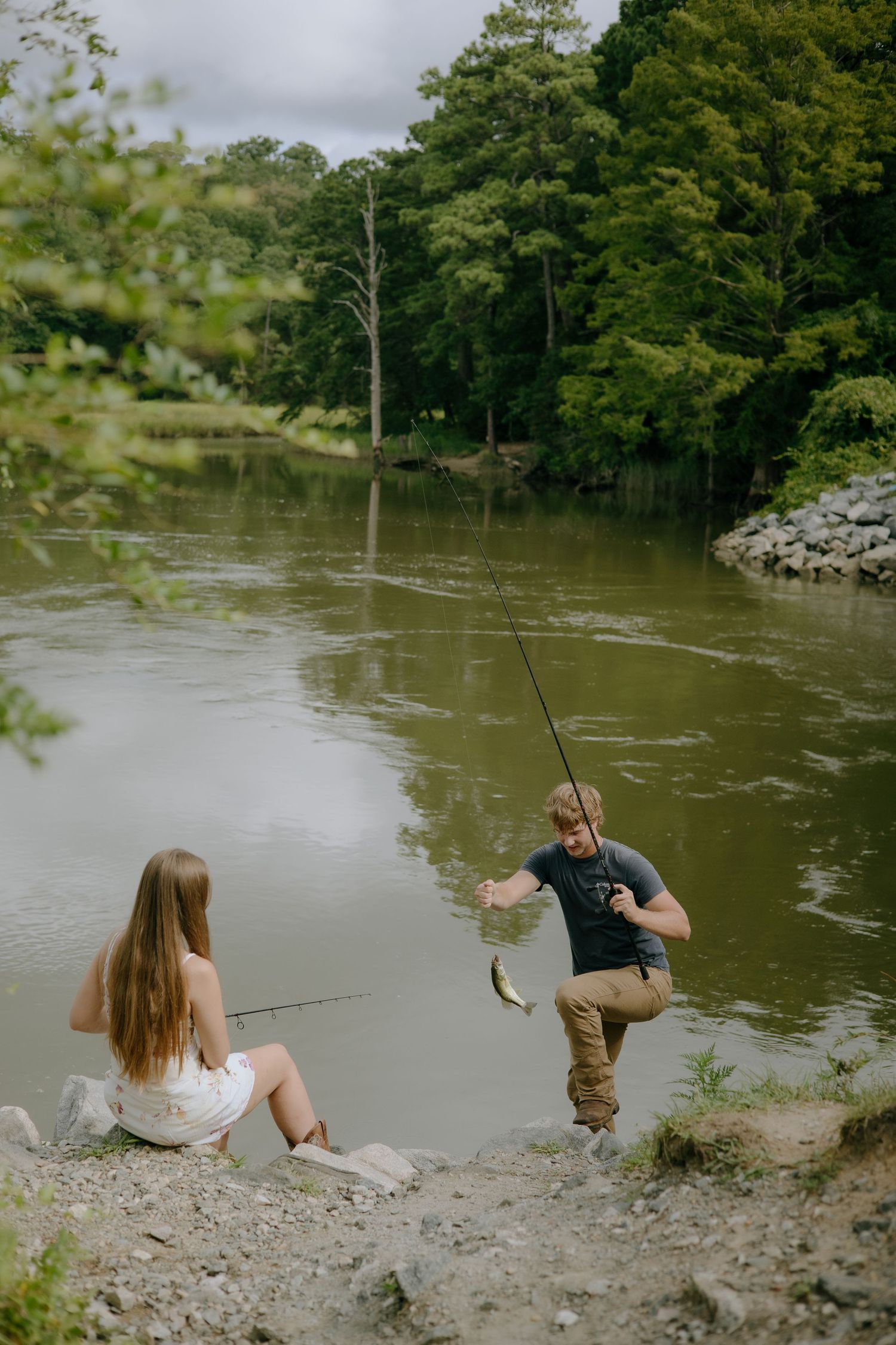 Two people relax and fish together on a rocky riverbank surrounded by lush greenery.