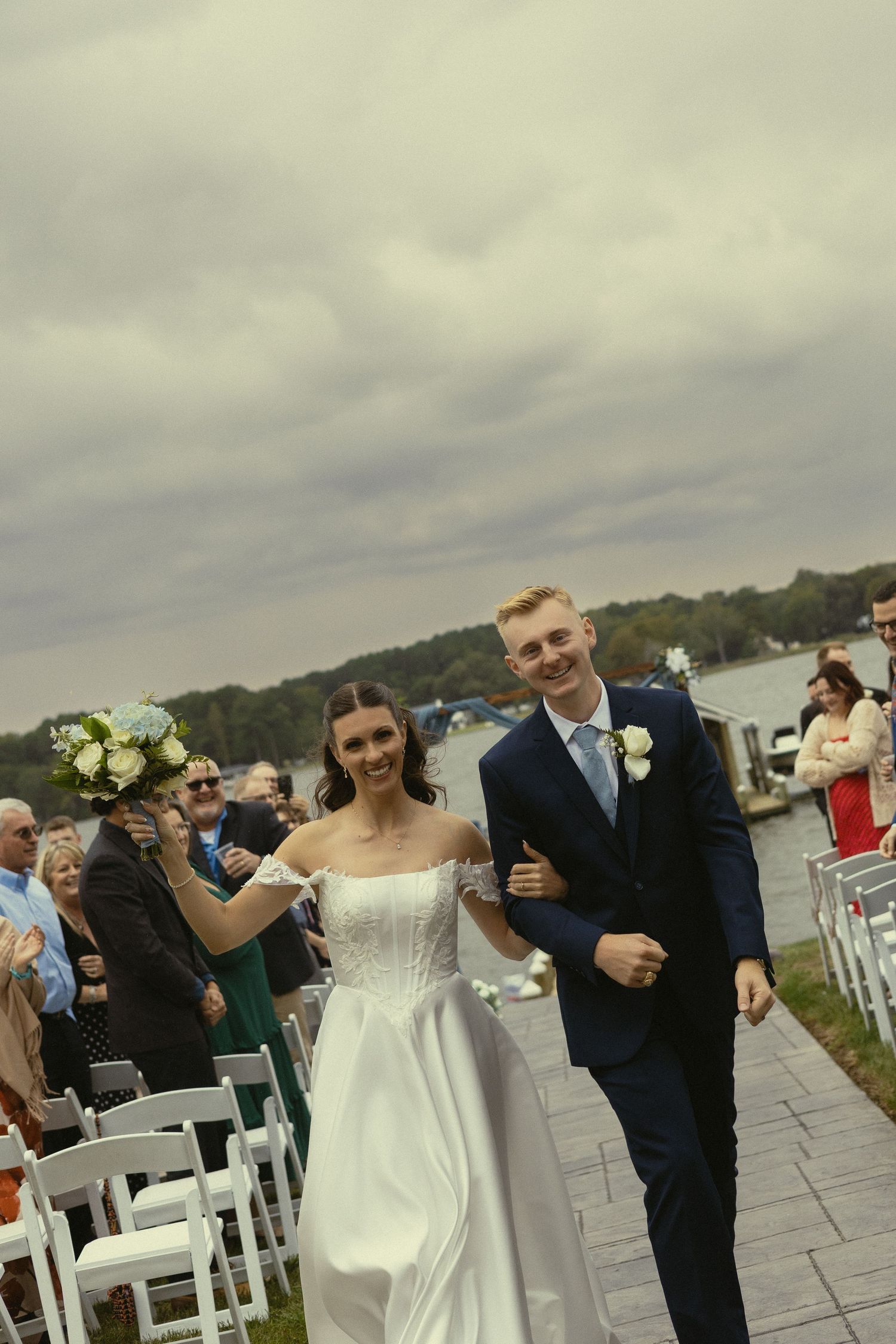 Joyful newlyweds walk down outdoor aisle by lakeside, bride in white gown holding bouquet, groom in navy suit.