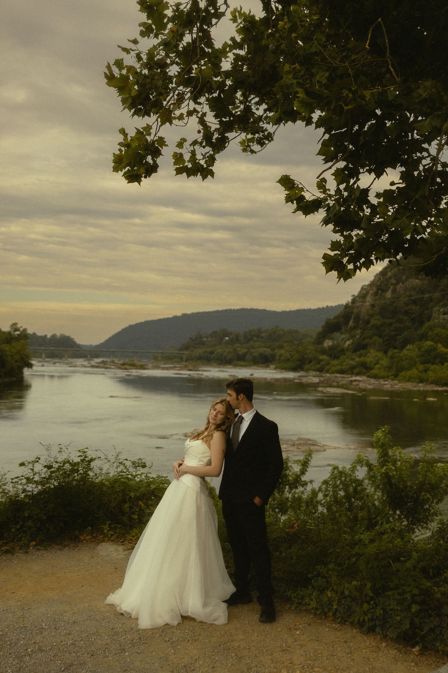 Bride and groom embracing by a scenic river with rolling hills and dramatic cloudy sky at dusk.