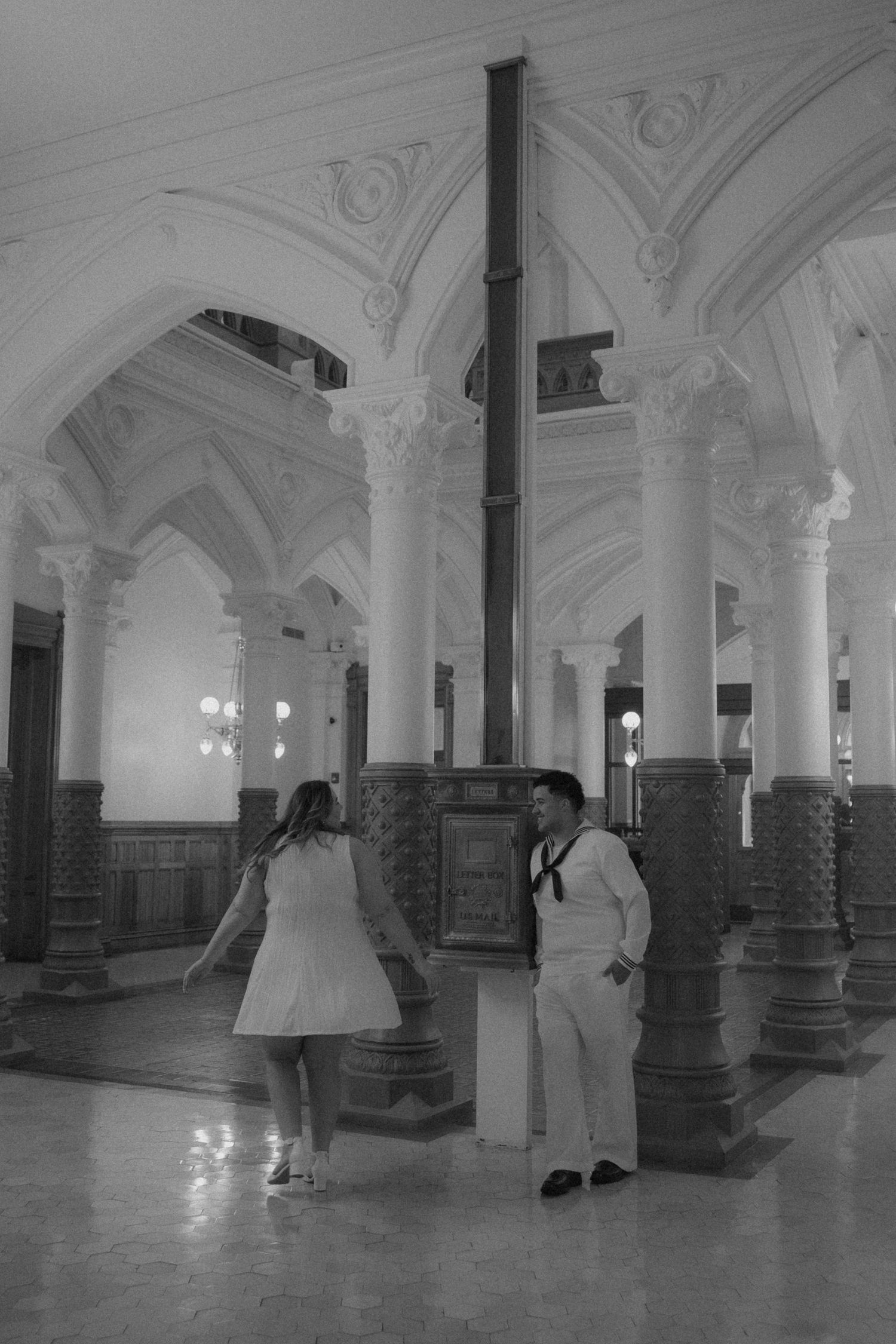 Black and white photo of a sailor in uniform leaning against a ornate column while a woman in a white dress approaches him.