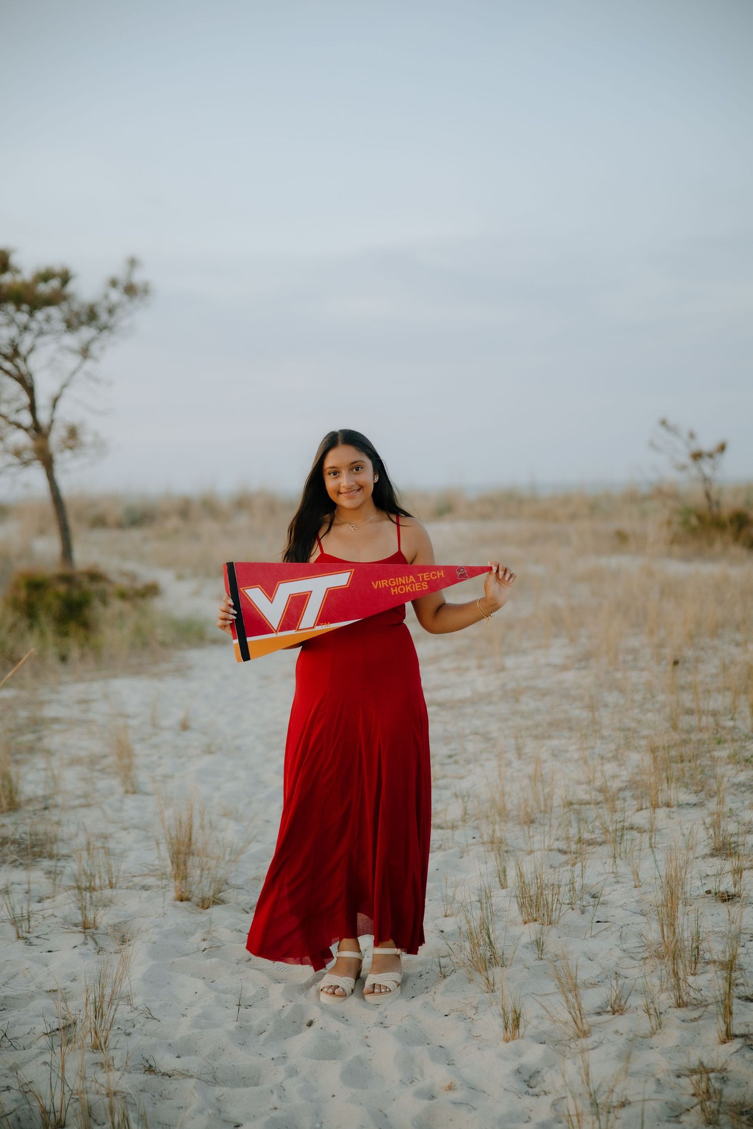 Young woman in red maxi dress holding Virginia Tech pennant on sandy beach path.