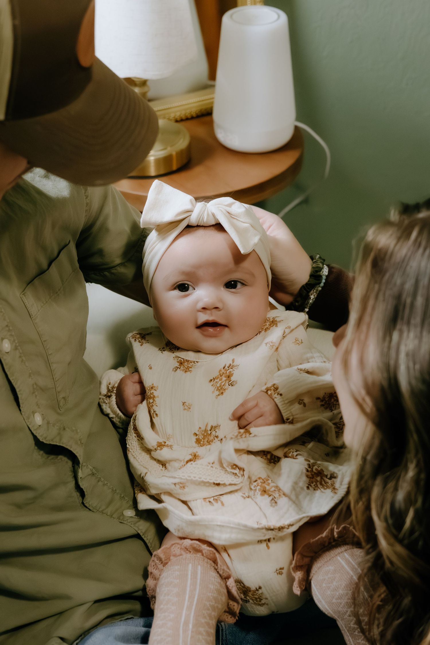 Baby in floral dress and bow smiling during playtime in a warmly lit room.