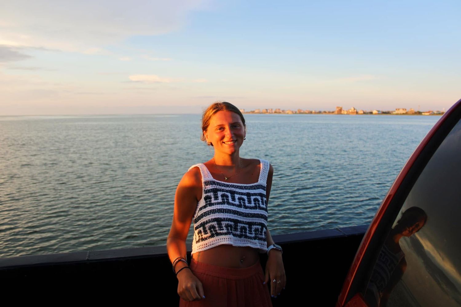 Woman smiling on a boat with ocean and city skyline at sunset in the background.