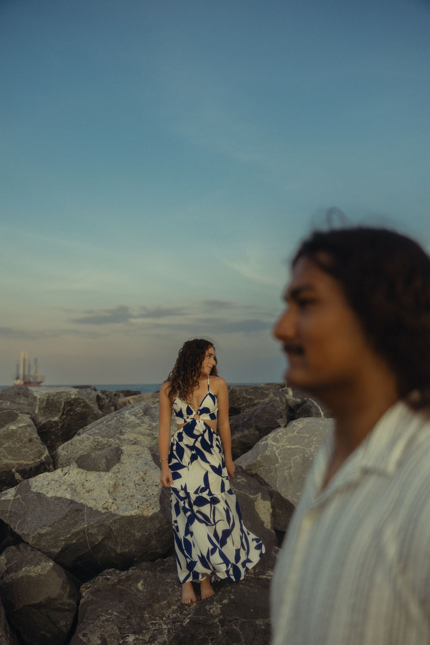 Woman in blue floral maxi dress stands on rocky shore at dusk, with blurred man in foreground.