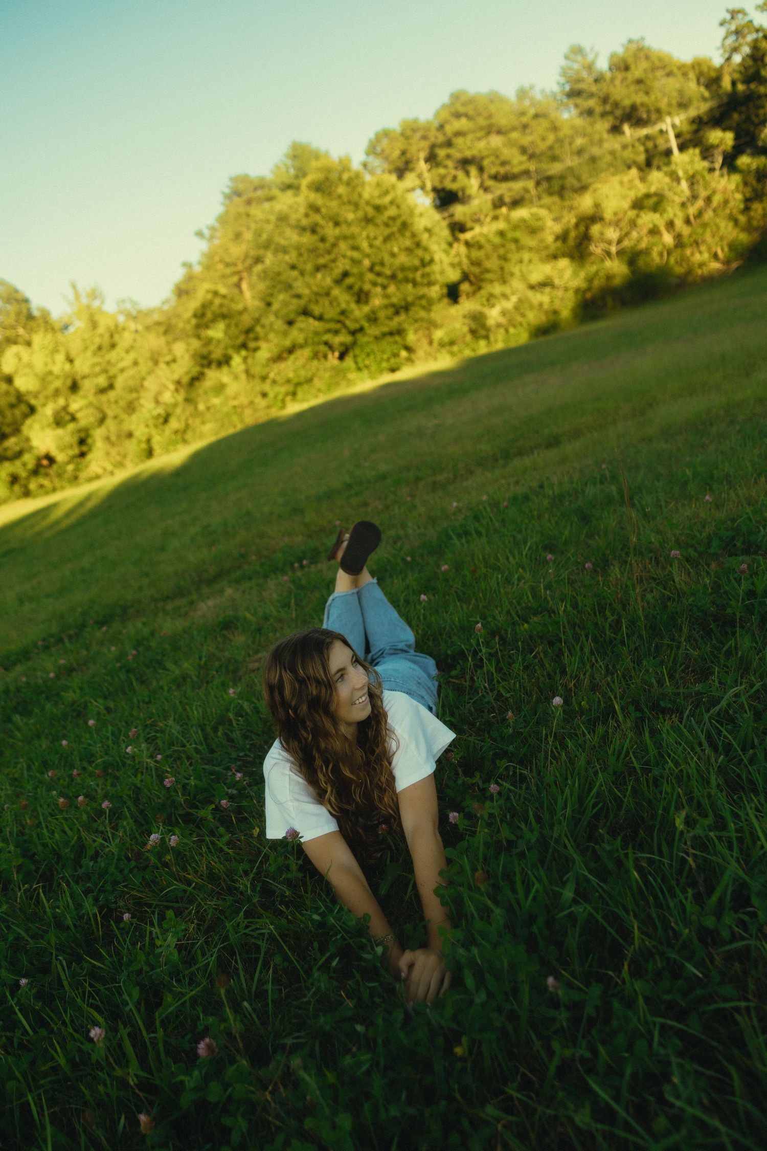 Young woman with long hair relaxing on lush green grass in a sunny park, wearing white top and jeans.