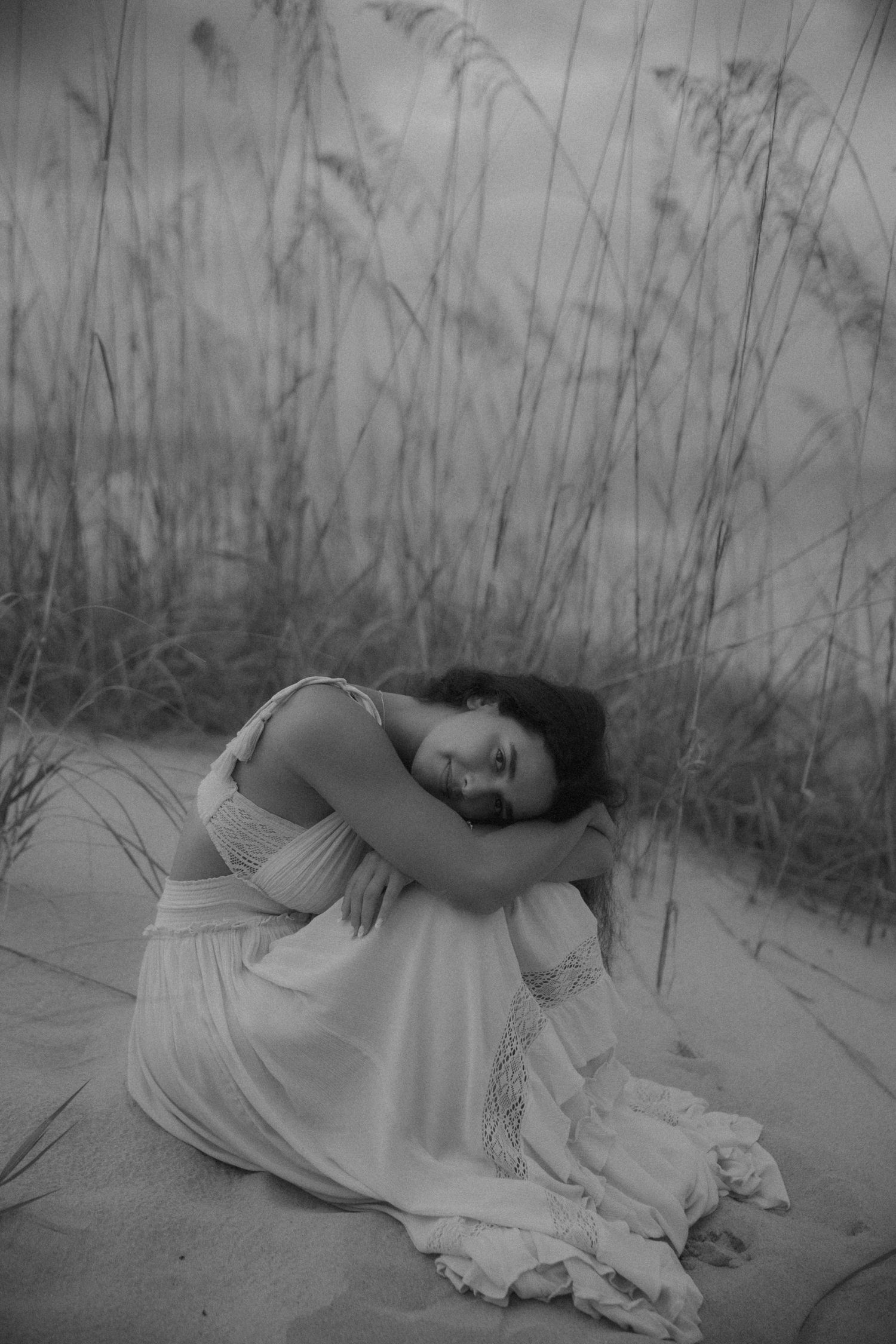Black and white photo of a woman in a white dress sitting on sand dunes surrounded by tall grass.