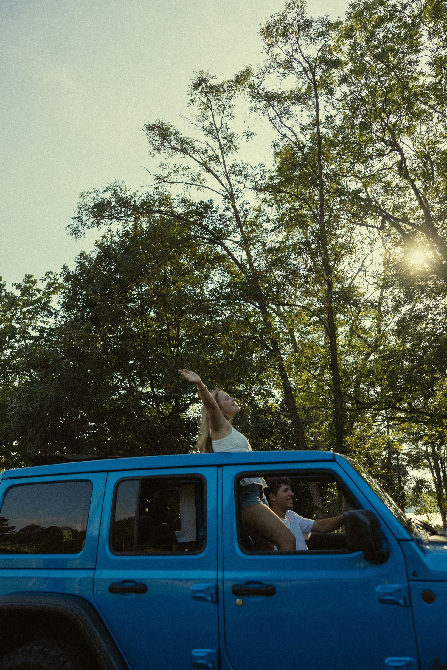 Couple enjoying a sunny day in a blue Jeep Wrangler, with one person standing through the open top surrounded by trees.