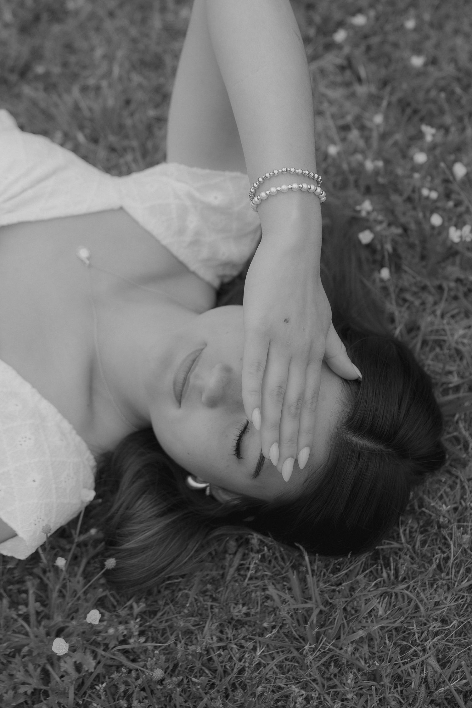 Black and white photo of a woman in white dress lying on grass, hand covering her face, wearing a pearl bracelet.