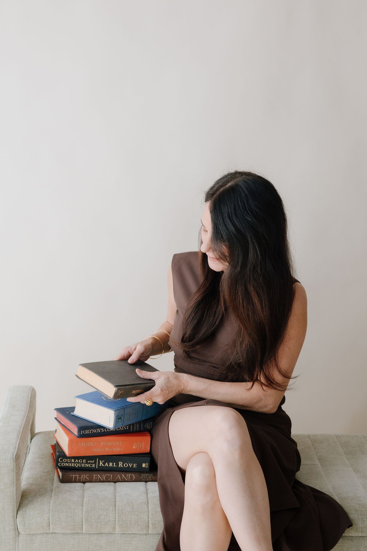 Woman in brown dress sits on a couch, browsing a stack of books against a neutral wall.