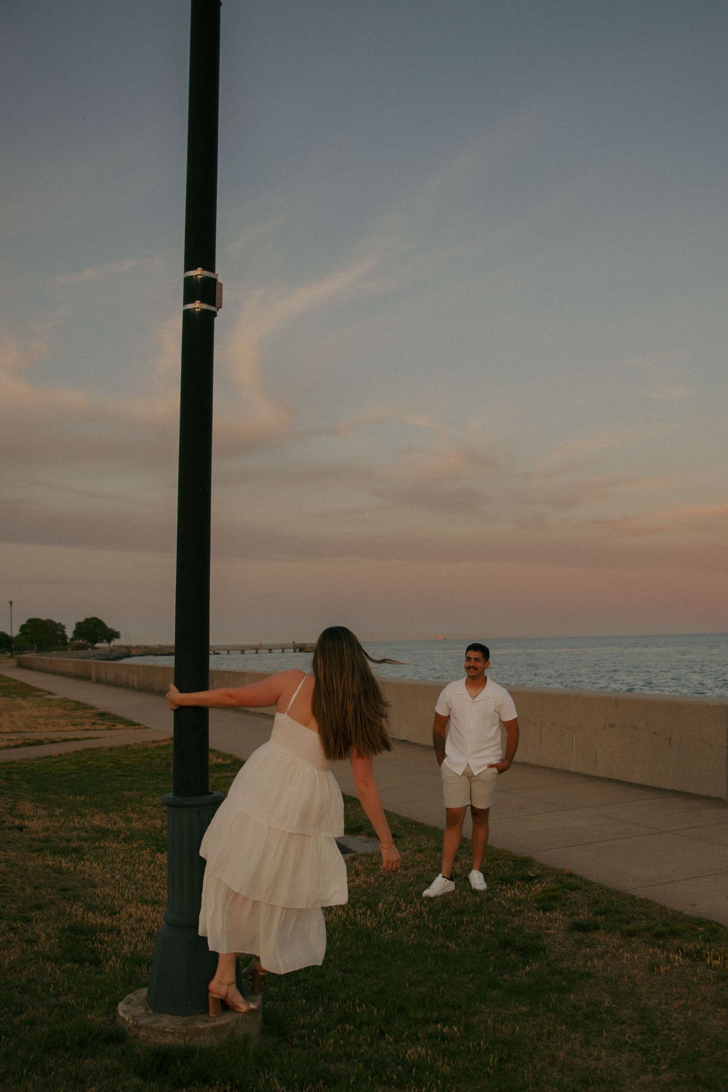 Couple in white outfits sharing a romantic moment by a lamppost near the ocean at sunset.