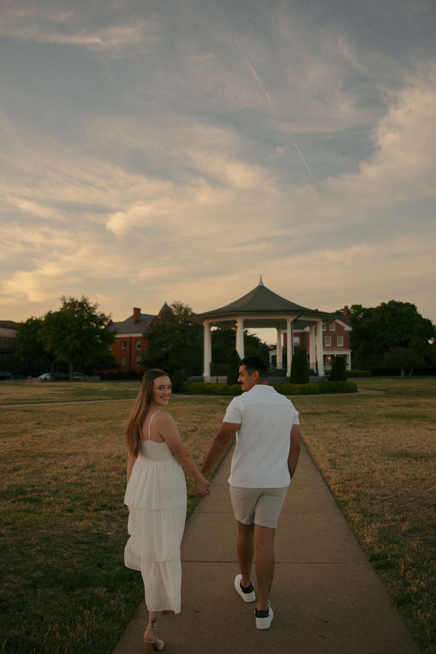 Couple holding hands walking toward a gazebo at sunset, woman in white dress looking back, golden hour sky.