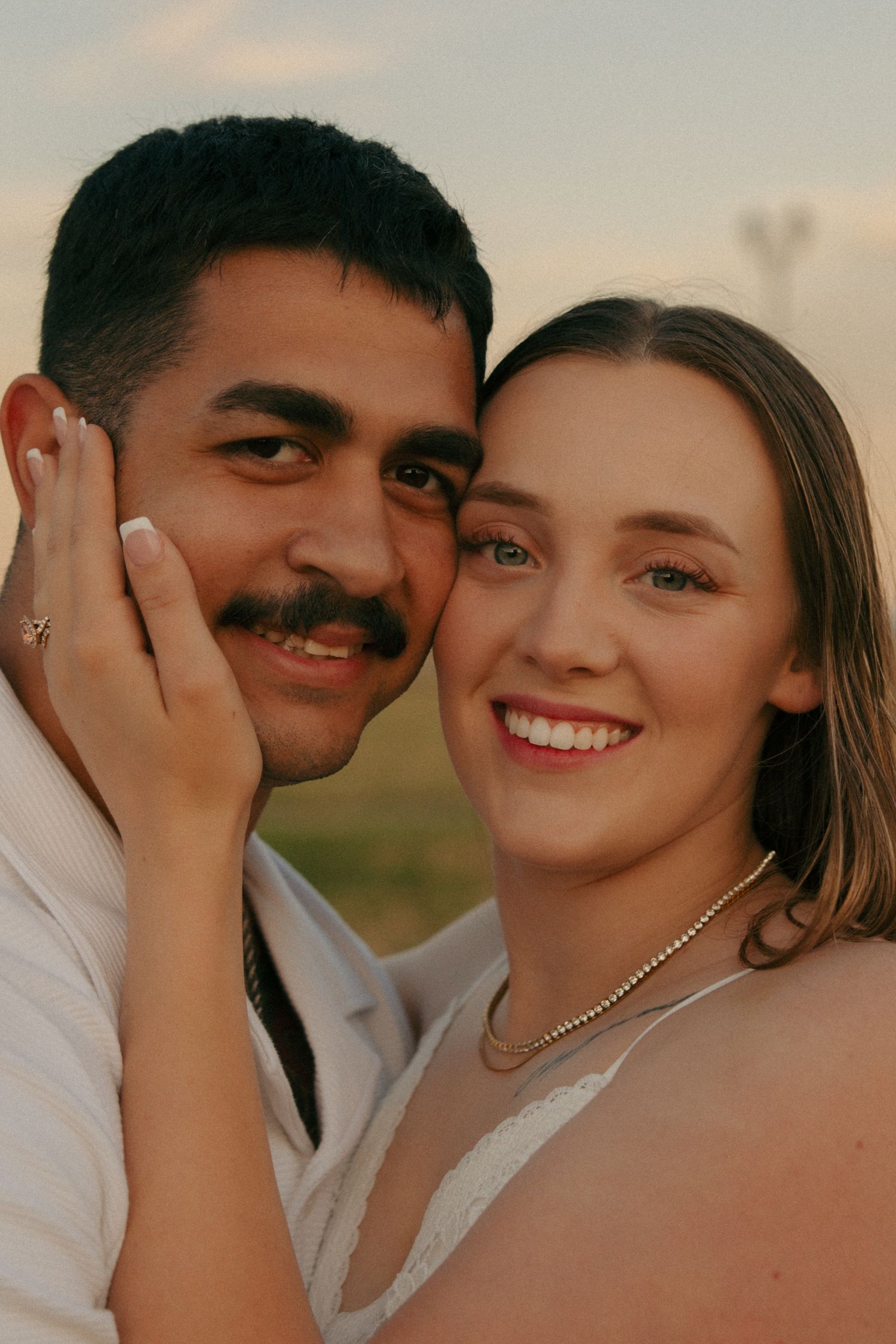 A smiling couple poses cheek to cheek at sunset, the woman wearing a pearl necklace and gently cradling the man's face.